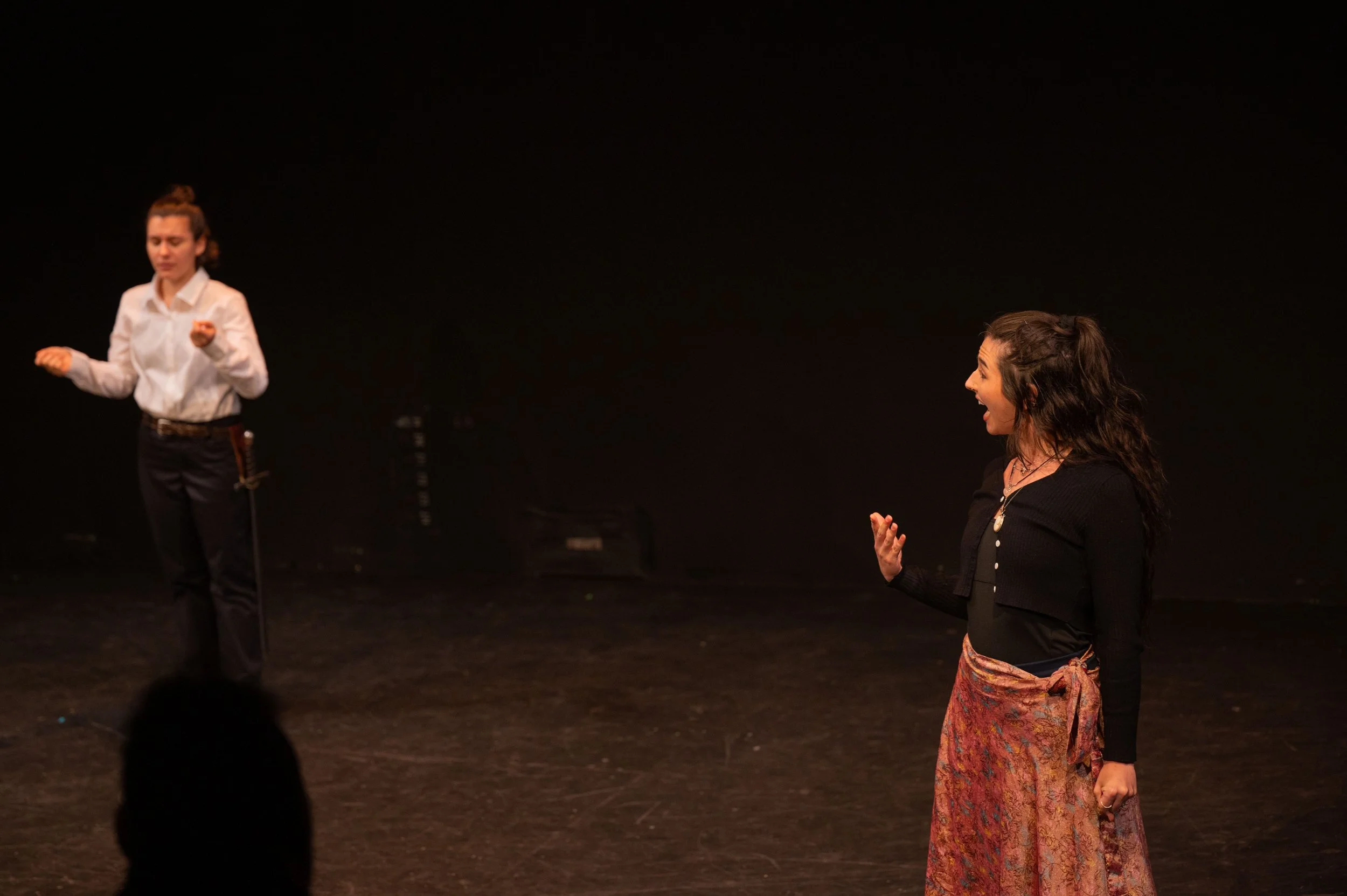 Two women are on a stage engaged in a theatrical performance or rehearsal. The woman on the right, dressed in a black top and a colorful patterned skirt, appears to be speaking or singing with an expressive gesture. The woman on the left, dressed in a white shirt and black pants, stands with her eyes closed, seeming to be listening or reacting to the other woman's performance. The stage is dark with minimal background.