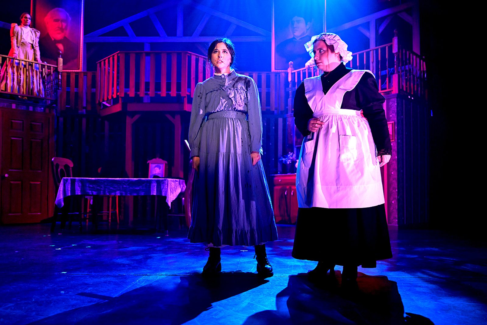 Two women in period costumes standing on a dimly lit stage with purple and blue lighting, set to resemble a historical interior with wooden balconies and framed portraits in the background.