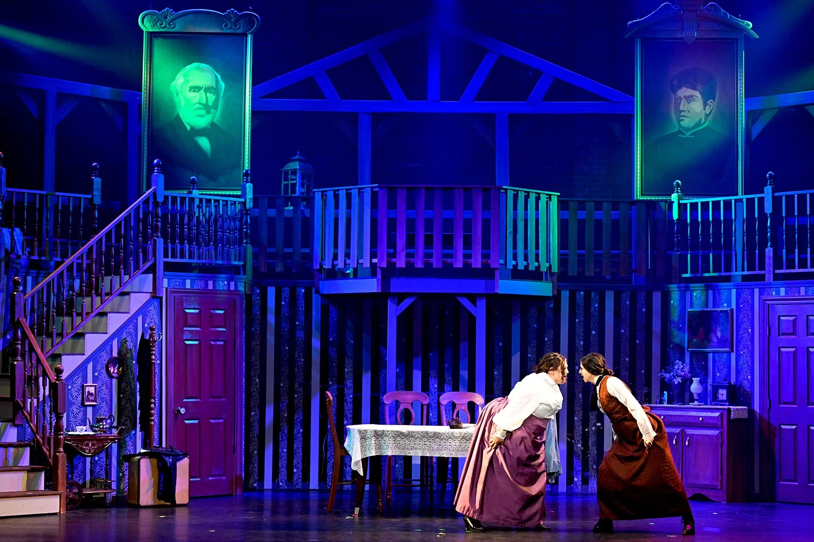 Stage scene from a theatrical play featuring two women in period costumes leaning toward each other in front of a large, colorful set resembling a house interior with portraits and staircase.