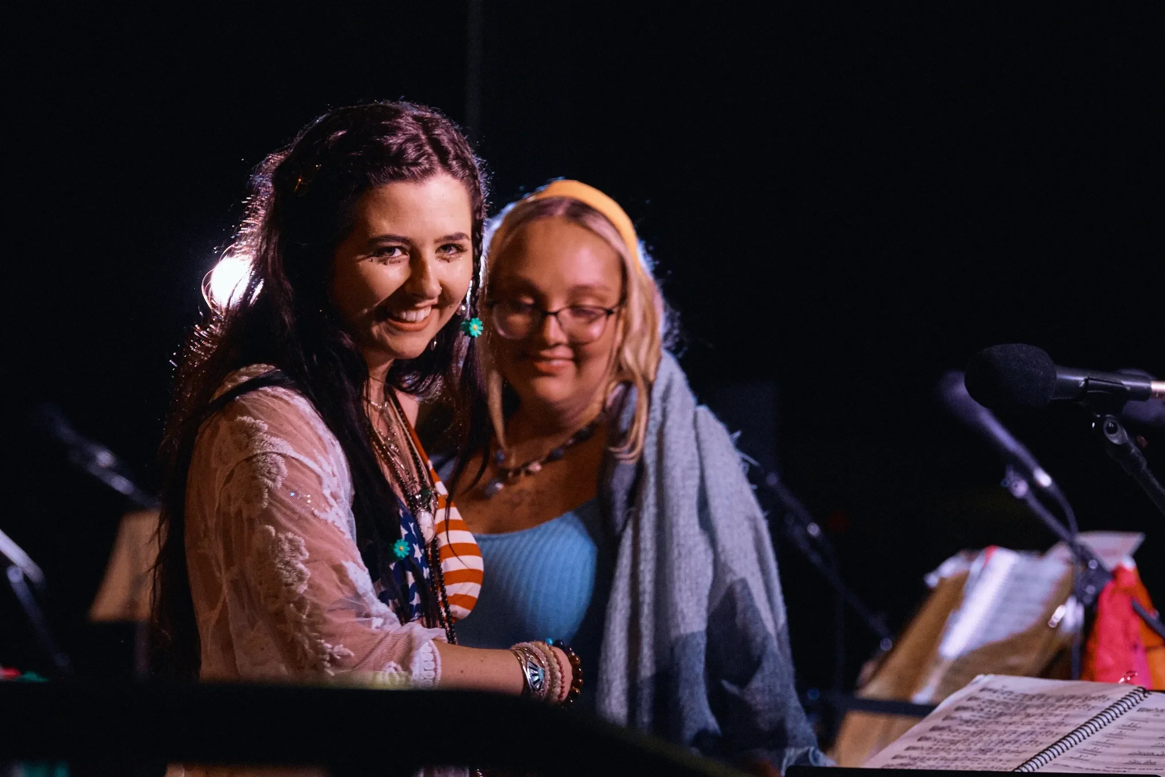 Two women smiling at a performance or event, with microphones and music sheets in front of them.