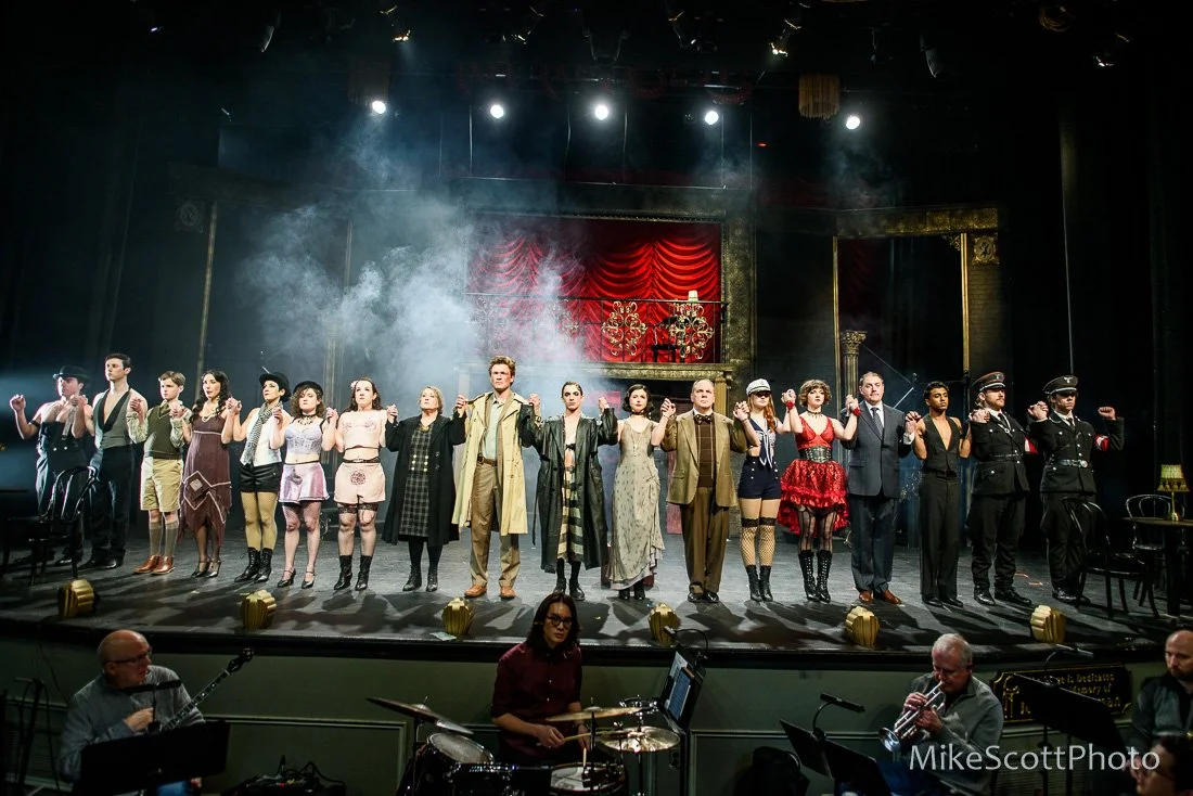 The cast of a theatrical production standing in a line on stage holding hands, with a red curtain and chandeliers in the background. Musicians are seen in the foreground.