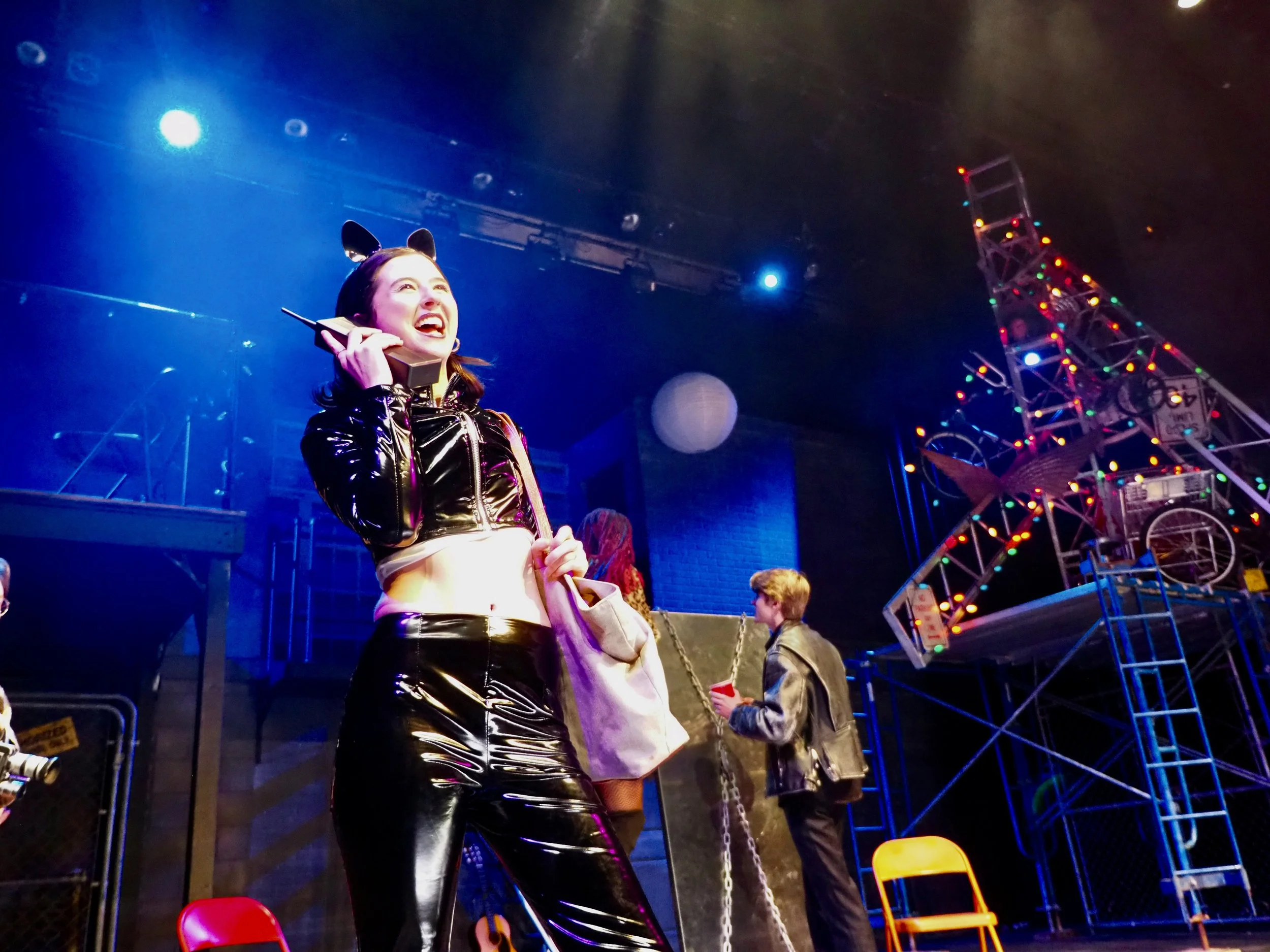 A woman dressed in a shiny black latex outfit with cat ears accessory, smiling and talking on a retro mobile phone at an indoor amusement park or carnival, with a ferris wheel decorated with colorful lights and a man in a leather jacket holding a red cup in the background.