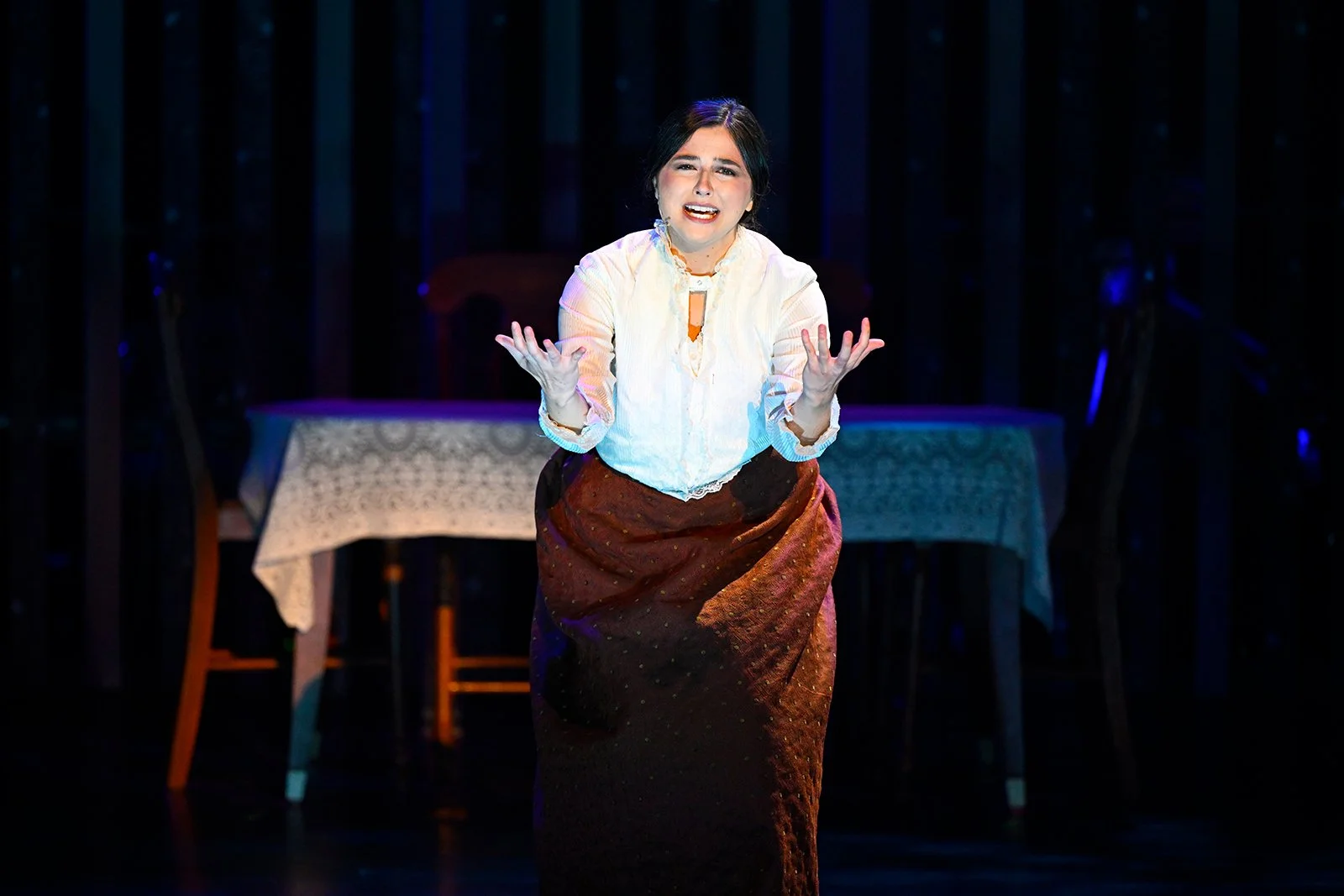 A woman on stage performing, wearing a white blouse and a long brown skirt, with a table and chairs in the background.
