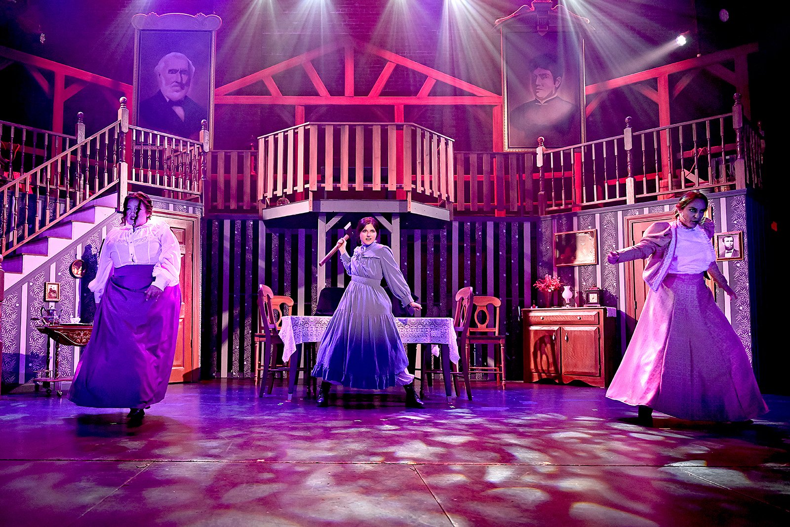 Stage scene from a theatrical play with three women in period costumes performing in a set resembling a vintage house interior, with portraits hanging on the wall and dramatic lighting.