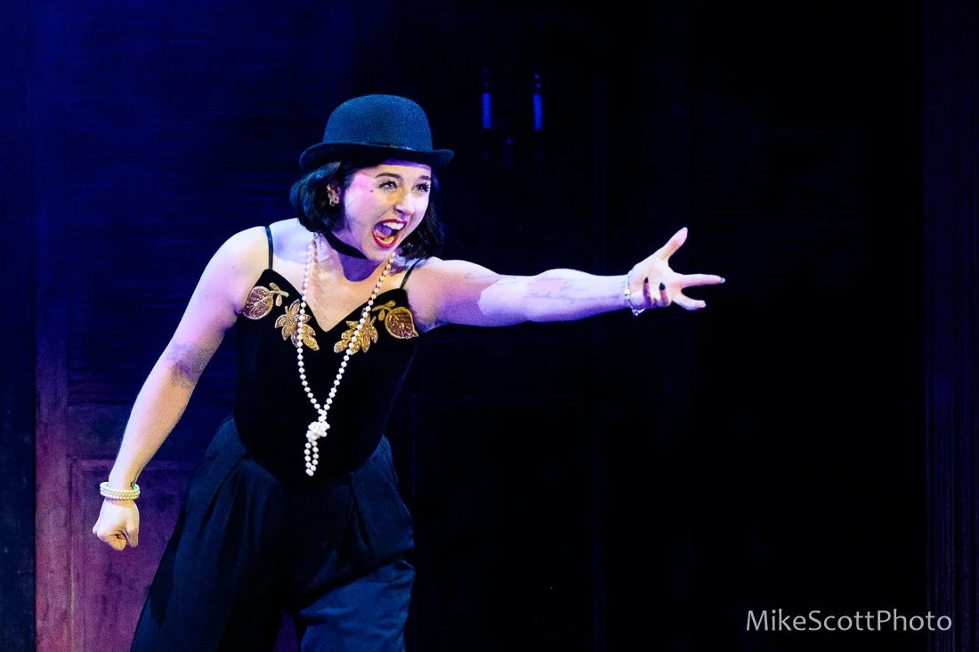 A woman wearing a black hat and dress, with jewelry, on stage, gesturing with excitement.