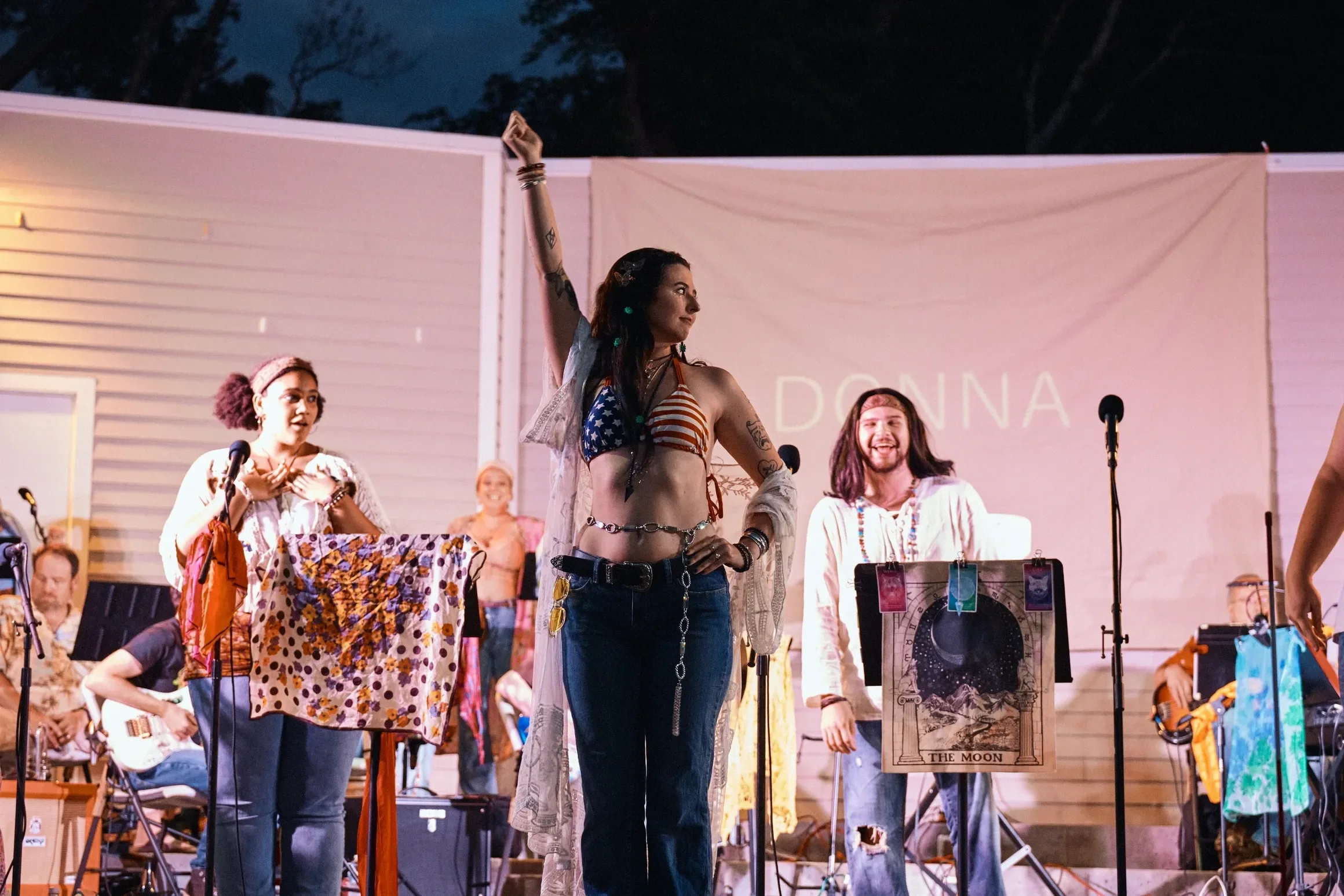 A woman wearing a stars and stripes bikini top, jeans, and a sheer cover-up stands on stage with her right arm raised. She has long dark hair with beads. Behind her are musicians and singers, some with guitars, performing before a large backdrop with the word "DONA."