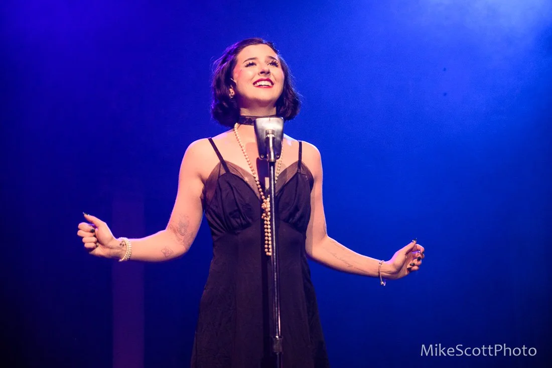 A woman singing on stage with a vintage microphone, wearing a black dress and jewelry, illuminated by colorful stage lights.