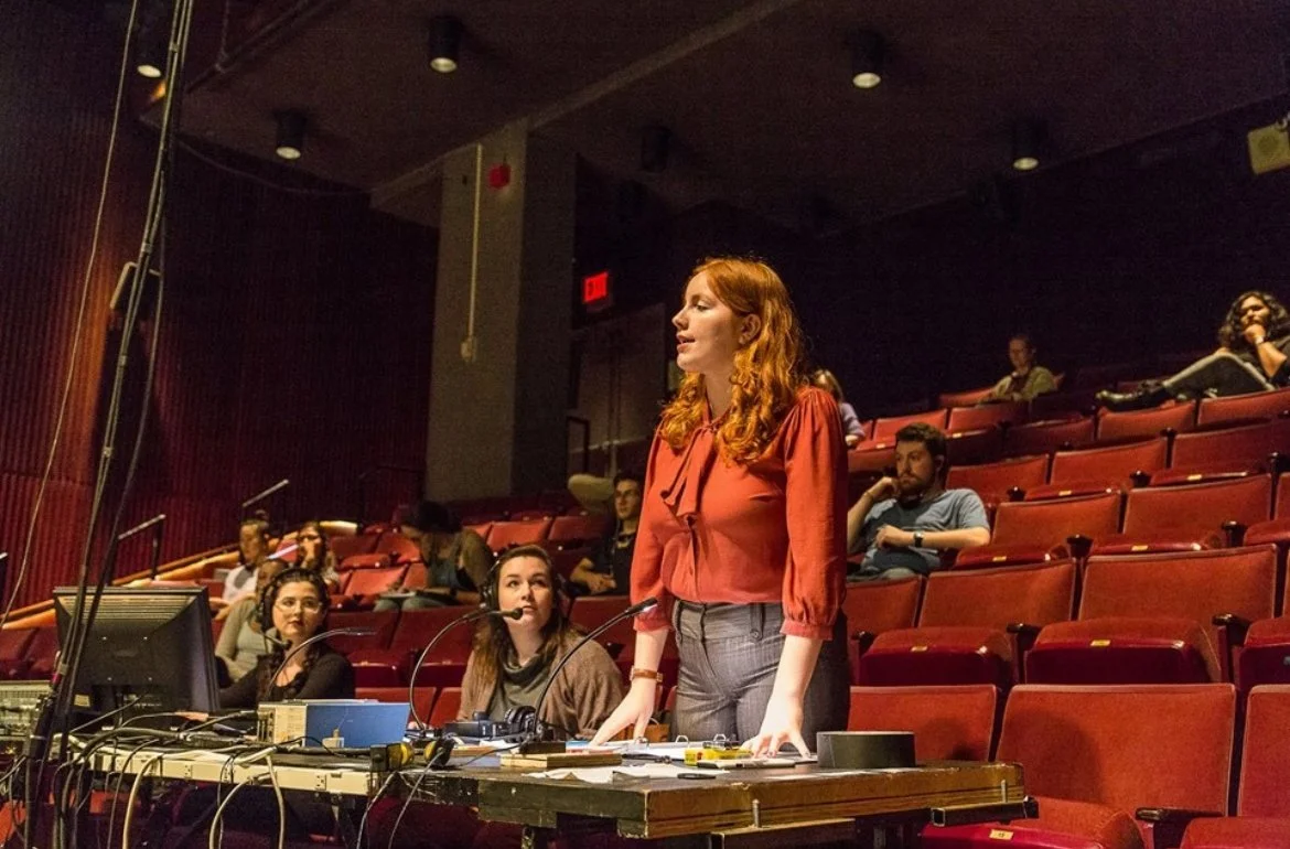 A woman with red hair standing at a table with microphones, while others sit behind her in an auditorium with red chairs.