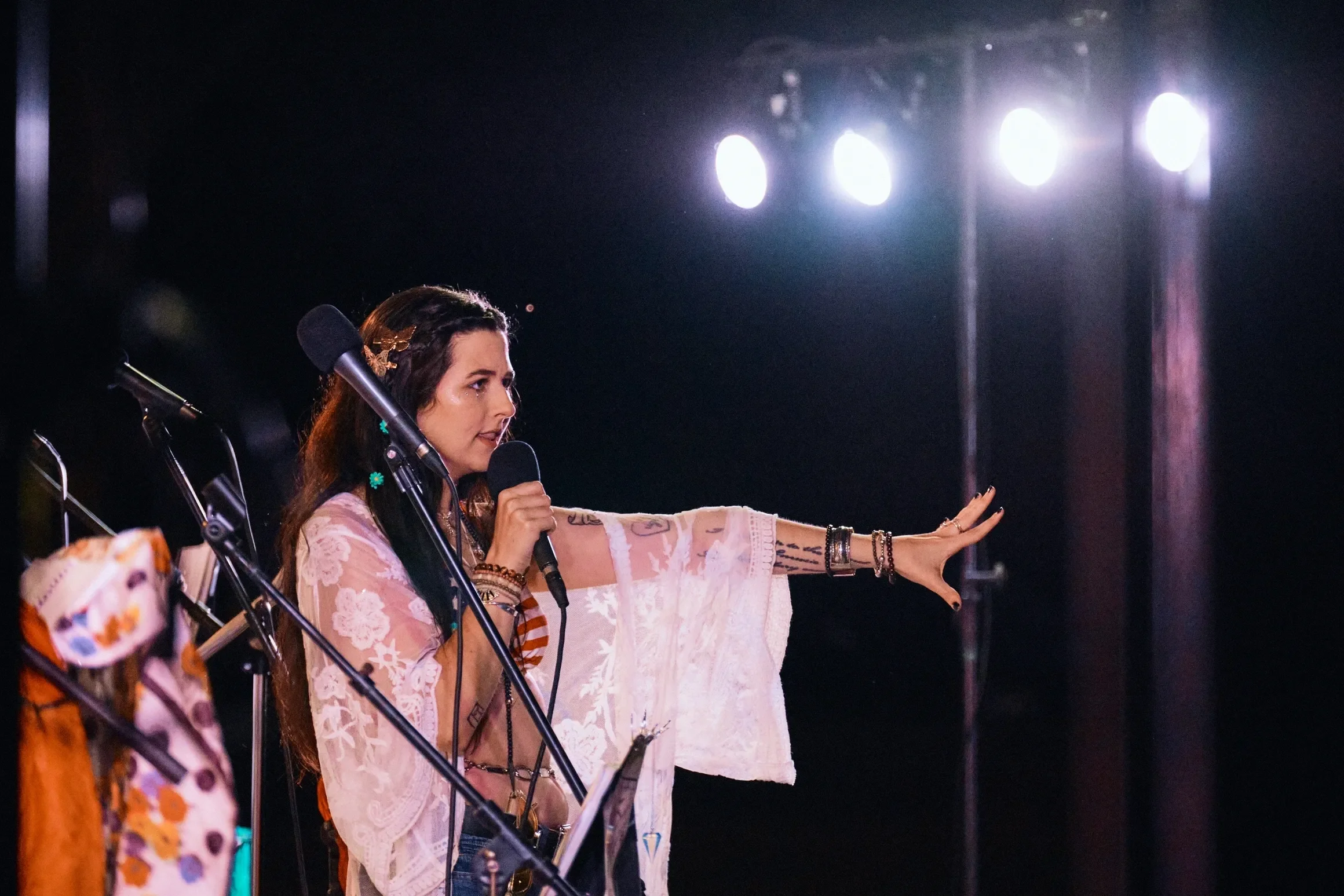 A woman with long dark hair and tattoos on her arm is speaking into a microphone on stage, with multiple microphones in front of her, and stage lights behind her.