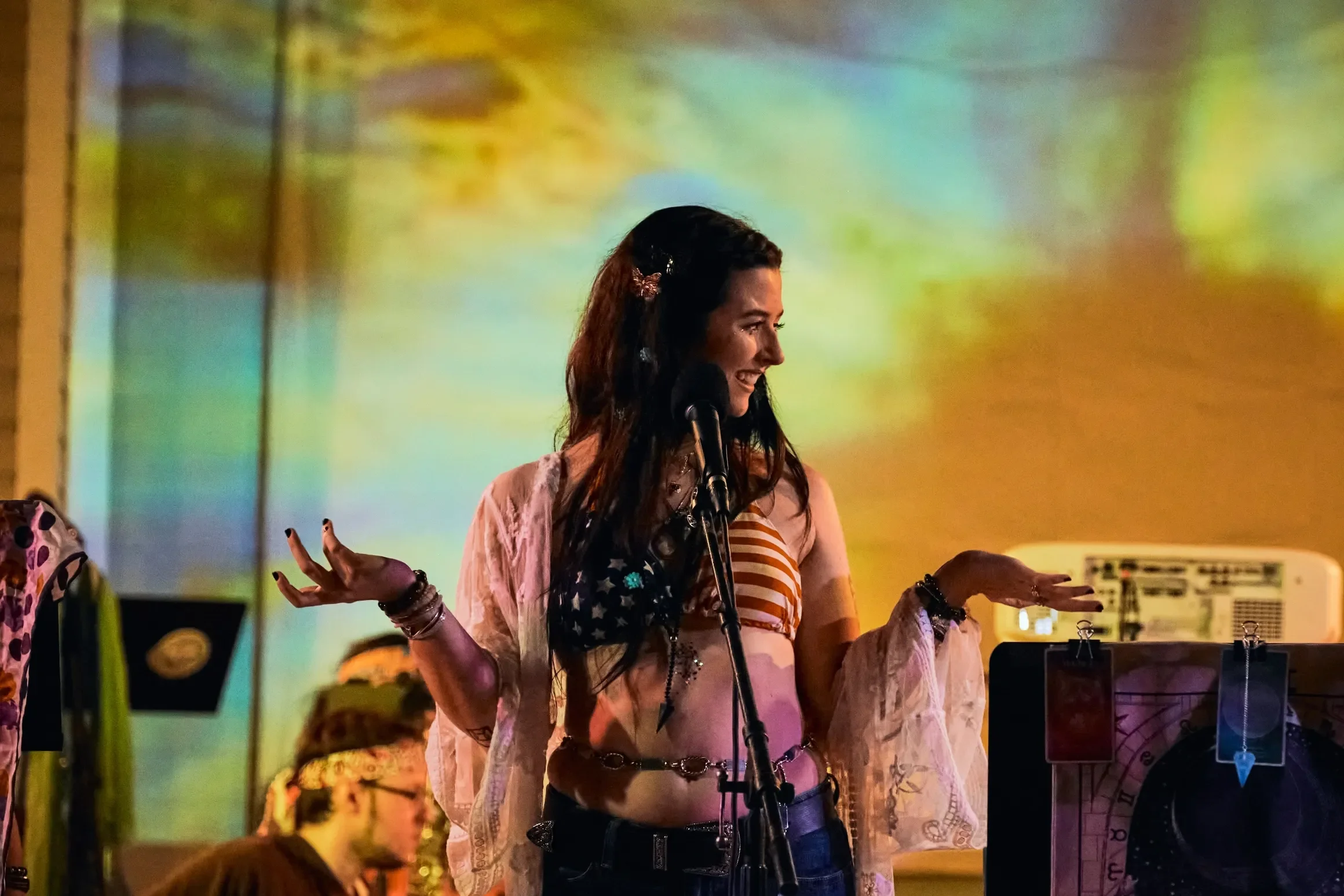 A woman with long dark hair, wearing a patriotic crop top with stars and stripes, and a sheer cover-up, standing in front of a microphone on stage, smiling and gesturing with her hands during a performance or event.