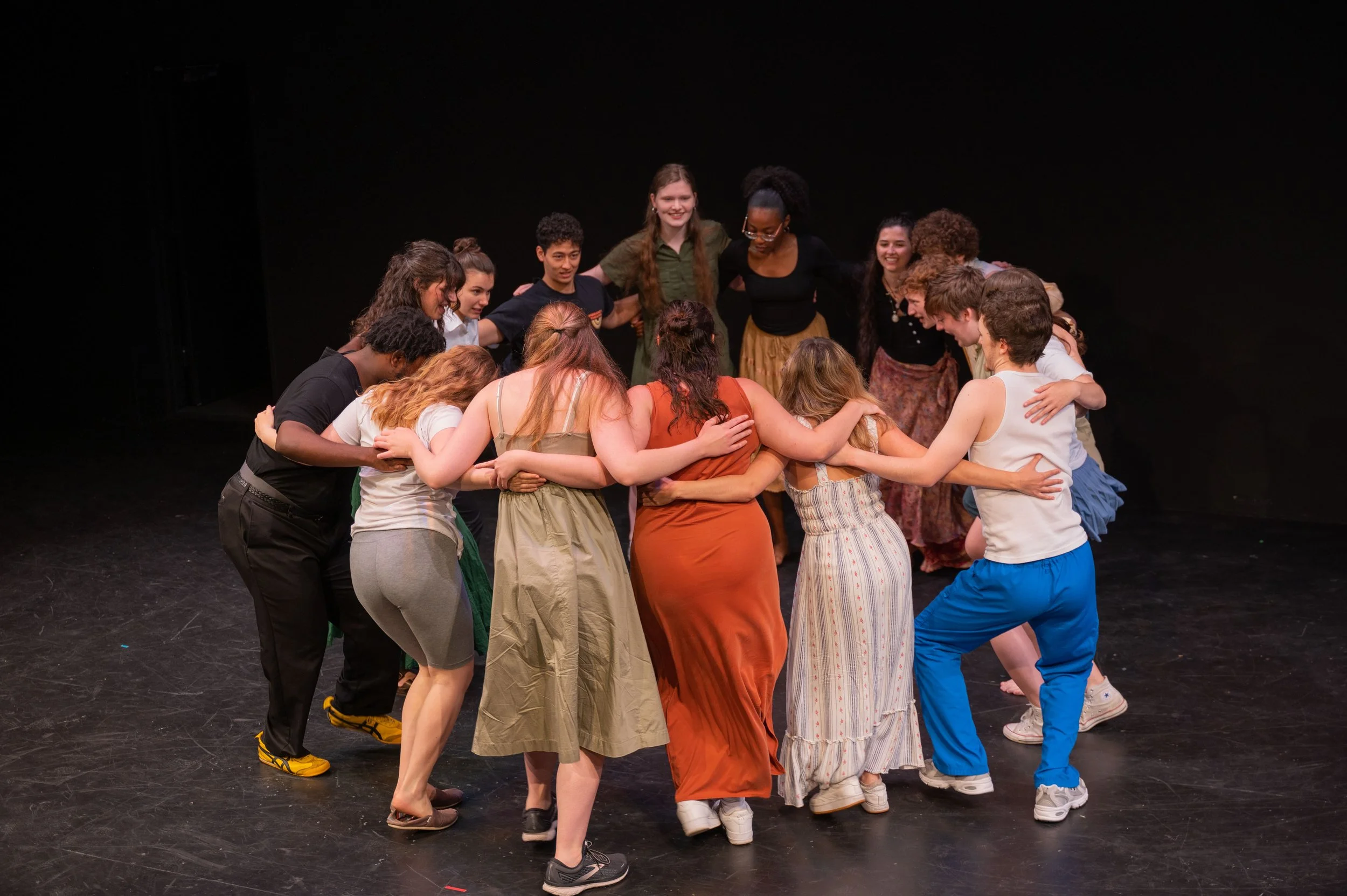 A diverse group of young people huddled together in a circle, smiling and embracing on a stage with a black background.