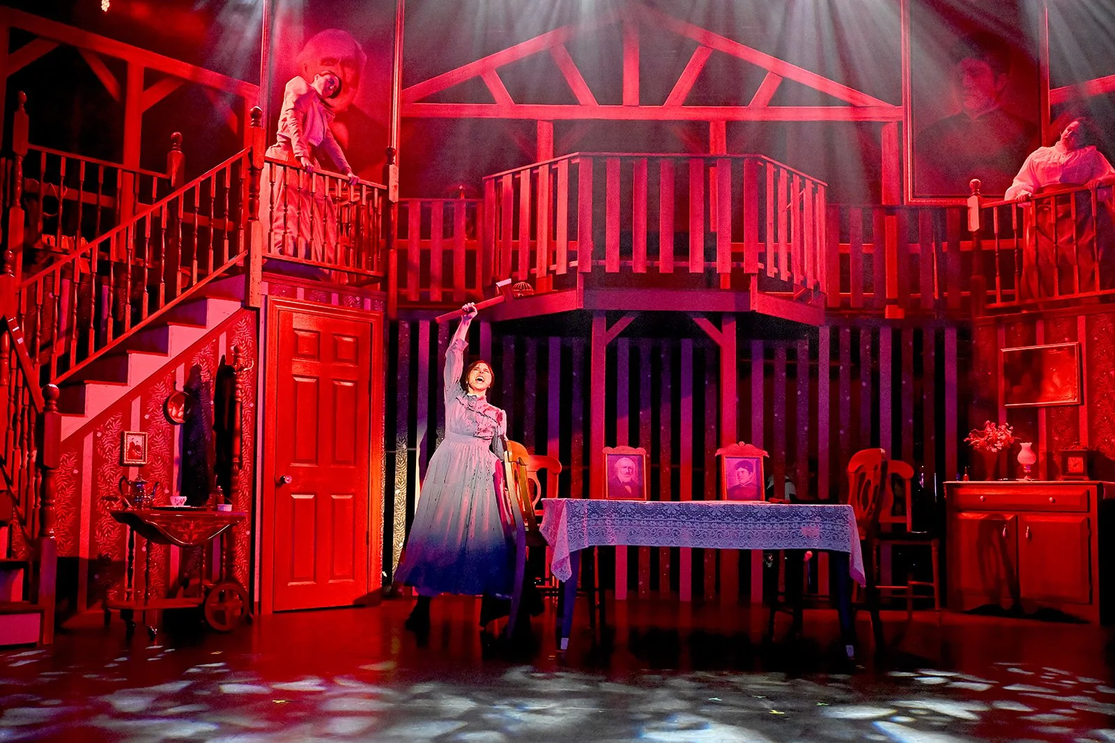 Stage scene with a woman in a vintage dress, raising a sword, surrounded by wooden furniture and portraits, with performers on a balcony, illuminated by red and purple lighting.