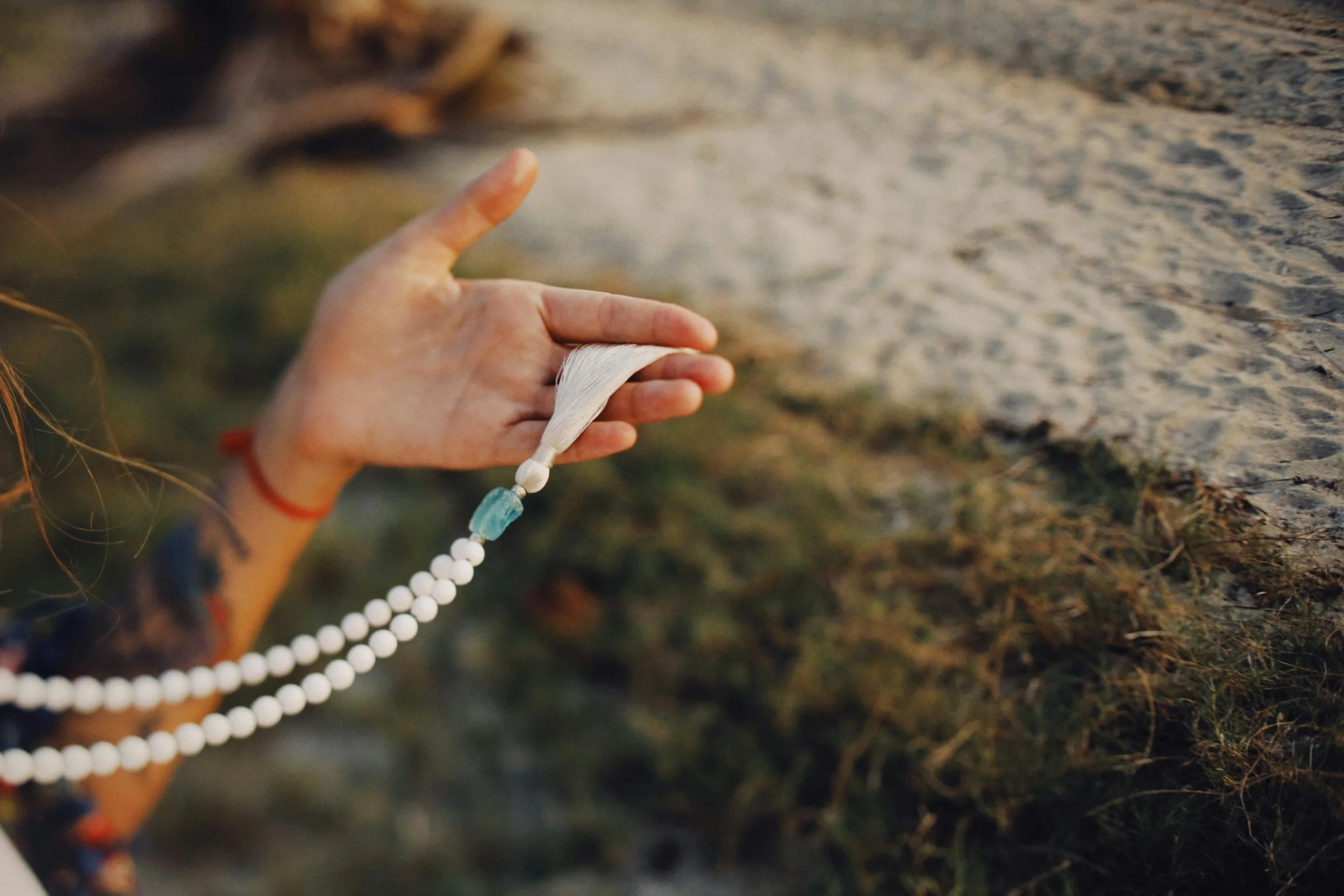 Close-up of a hand holding a strand of white mala beads with a tassel, outdoors with a blurred natural background.