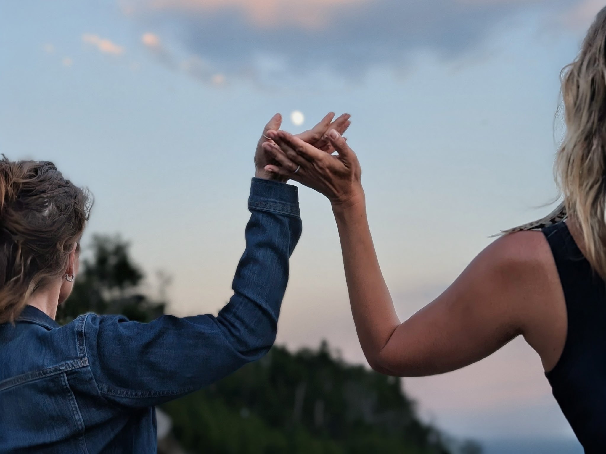 Two people raise their hands toward each other in a gentle touch or high-five against a soft evening sky