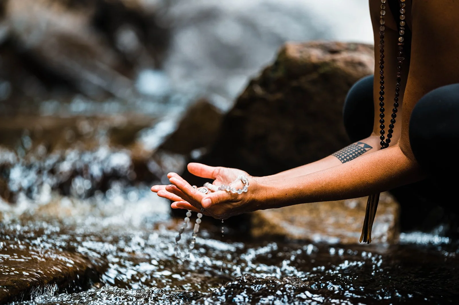 A person’s outstretched hand catches and releases water in a shallow, rocky stream; droplets fall between their fingers, and a small geometric tattoo is visible on their forearm.