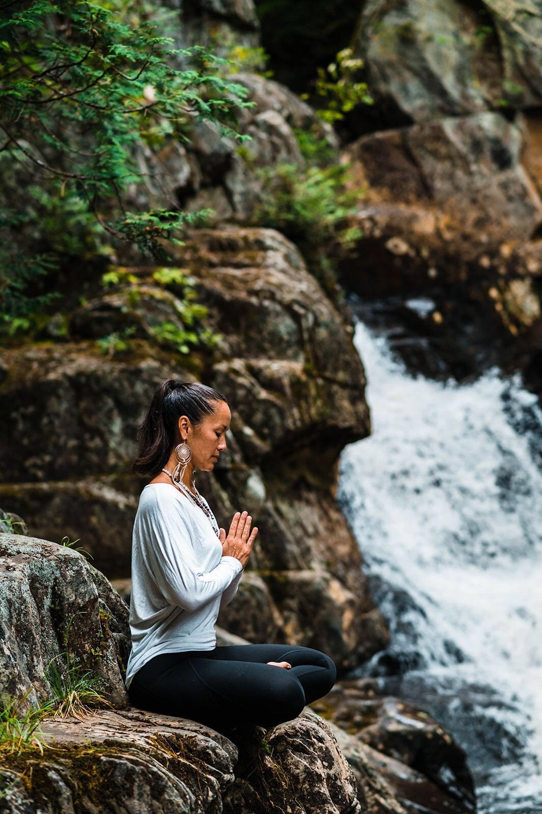 Steph while sitting on rocks beside a small waterfall, eyes closed and hands together in a meditative pose, surrounded by lush greenery.