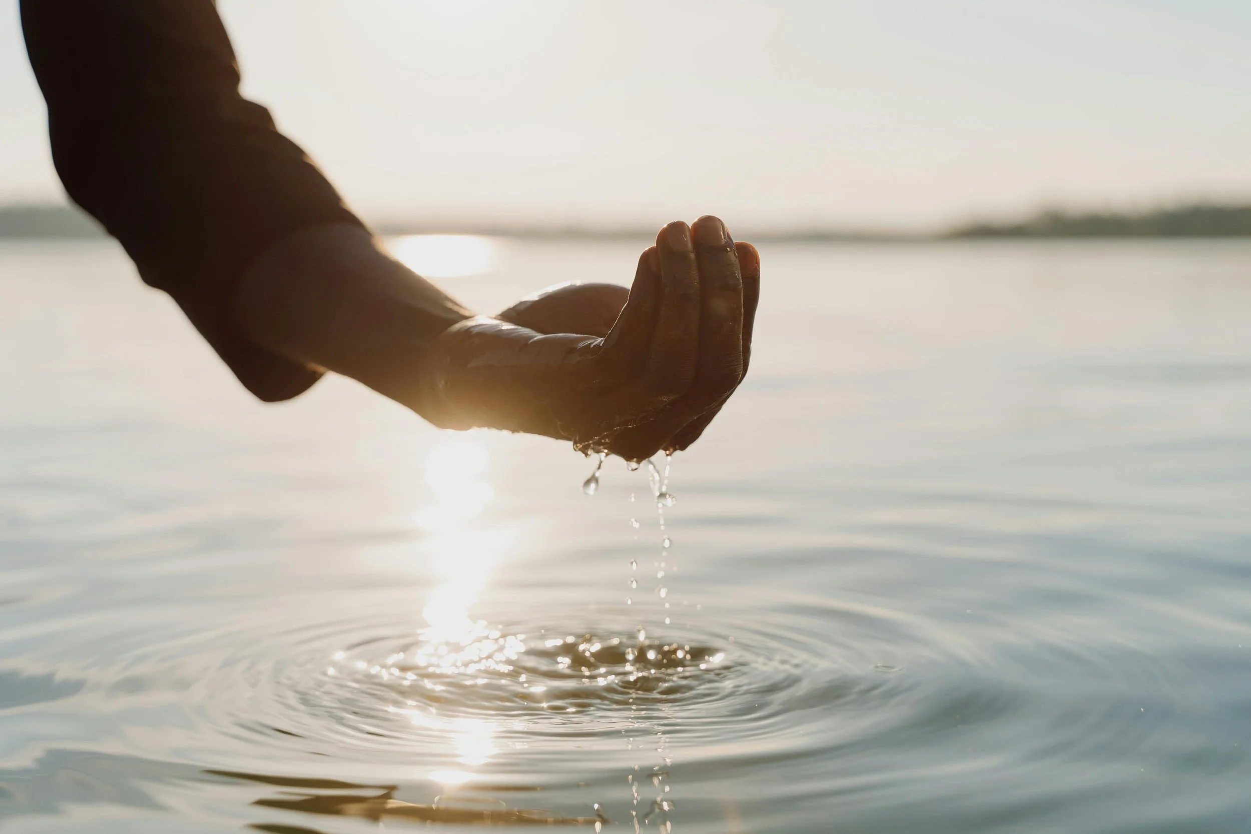 Hand cupping water over a calm body of water at sunset, with ripples forming below.