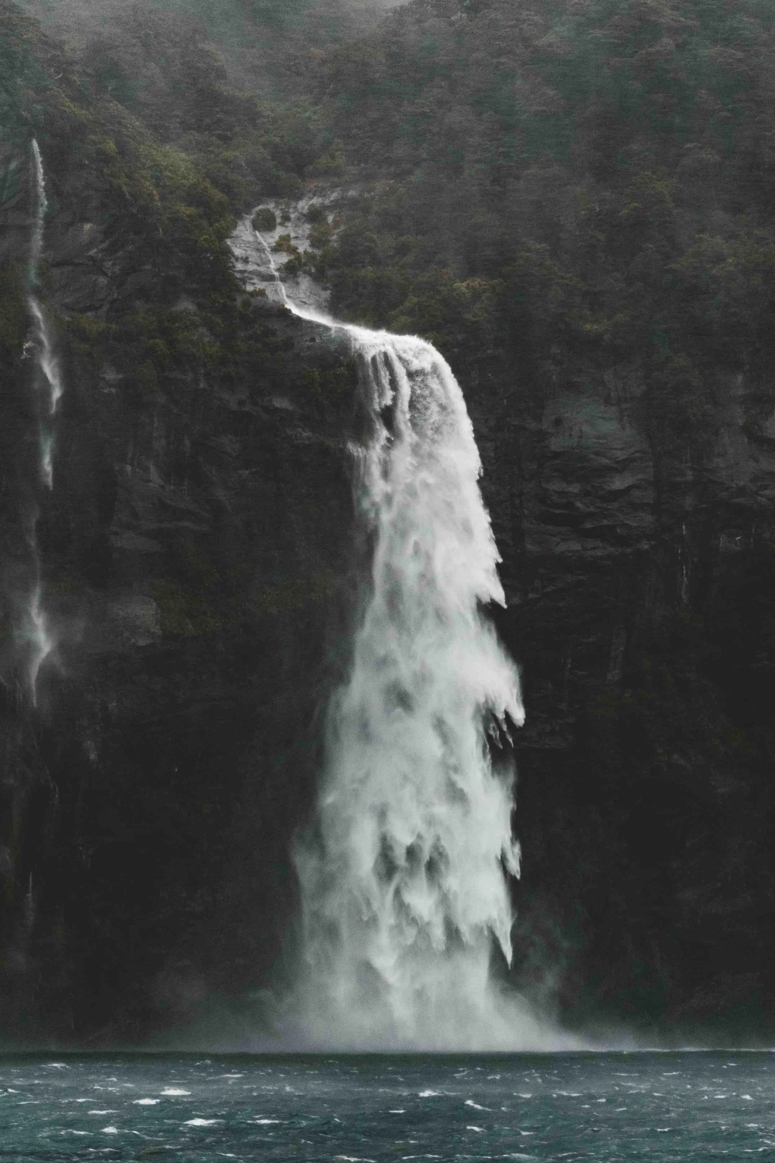 Tall waterfall cascading down a rocky cliff surrounded by mist and lush greenery.