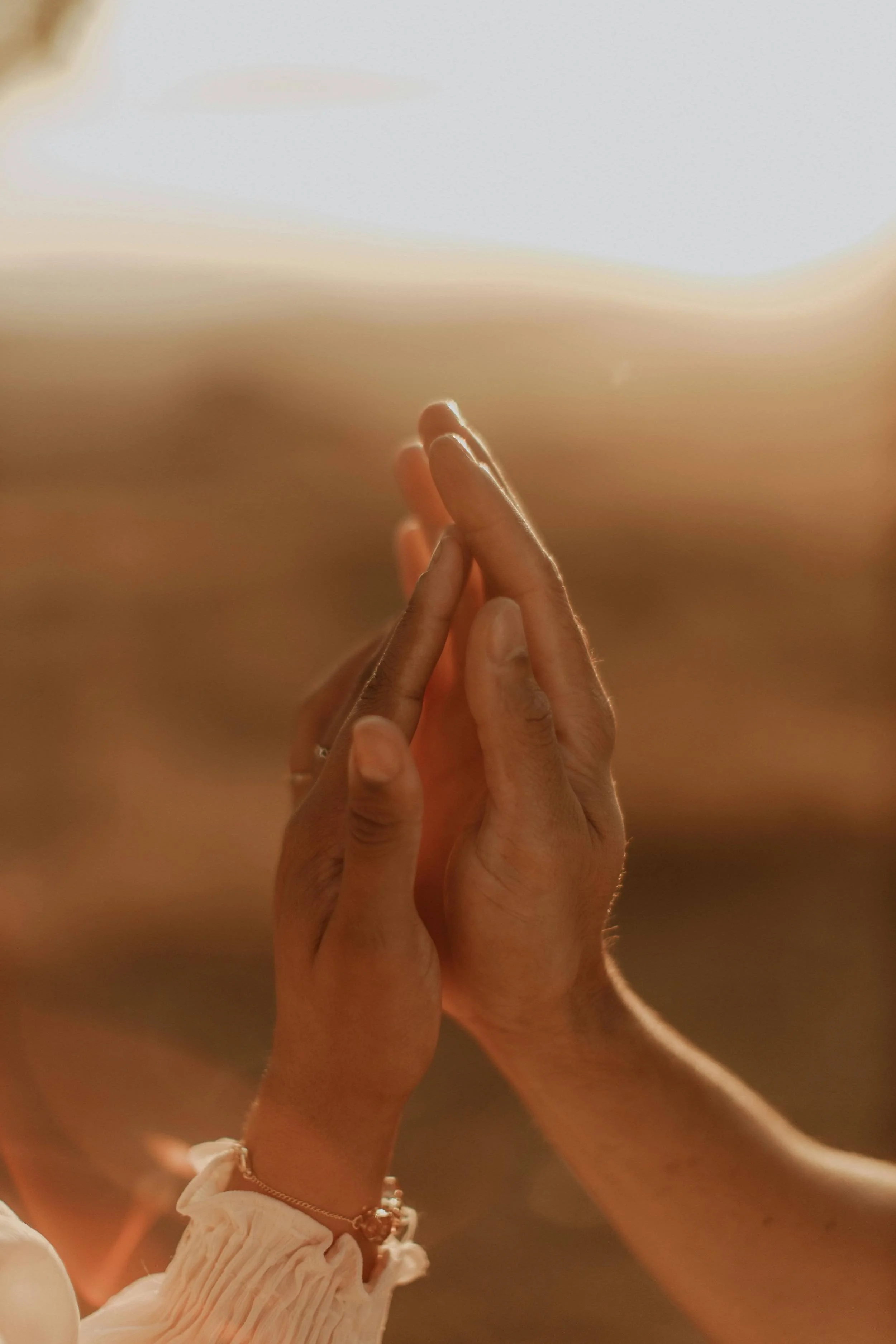 Two hands pressed together in a prayer-like gesture against a warm, sunlit landscape.