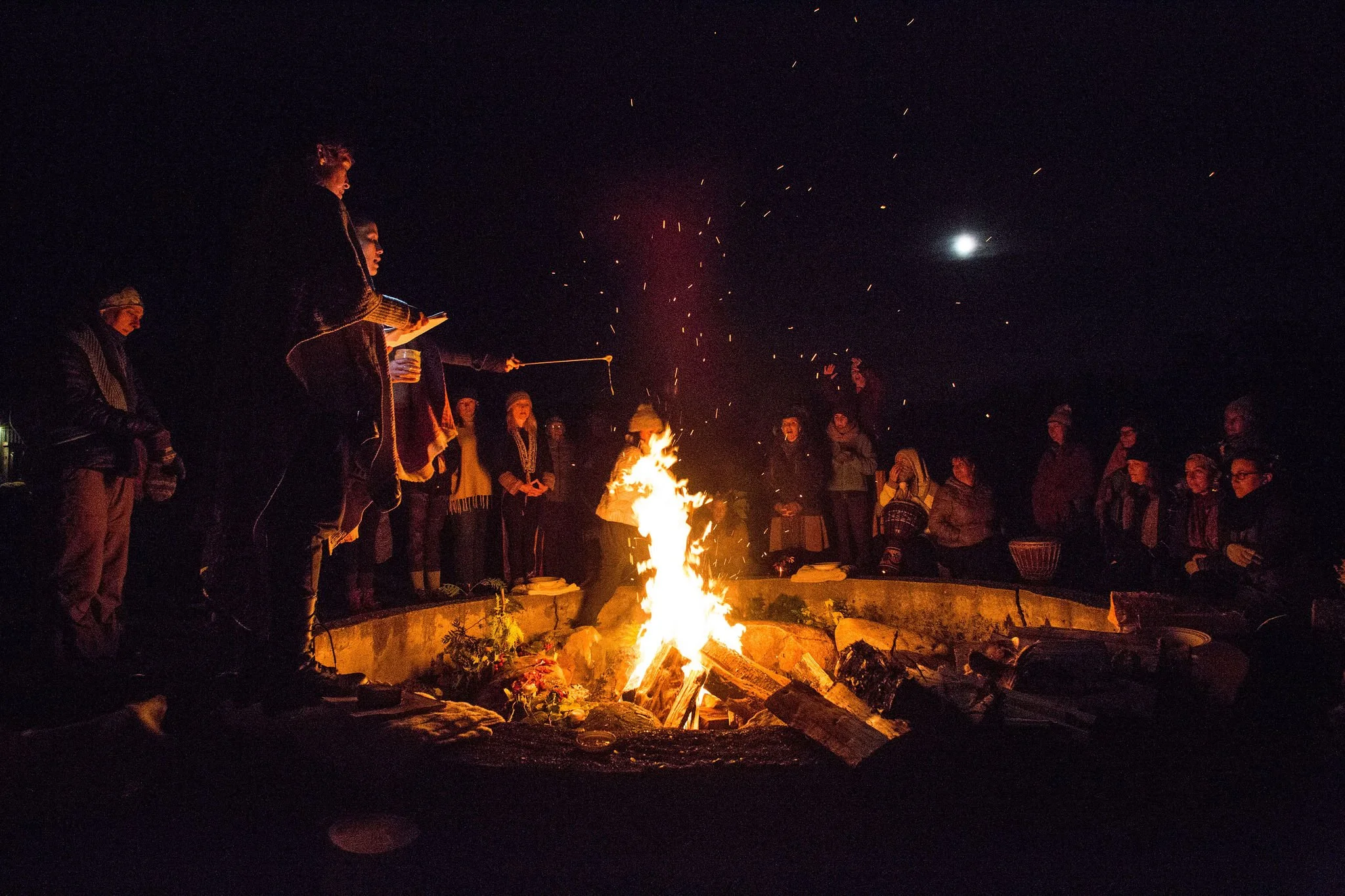 Group of people sitting around a campfire at night, with flames glowing against the dark surroundings.