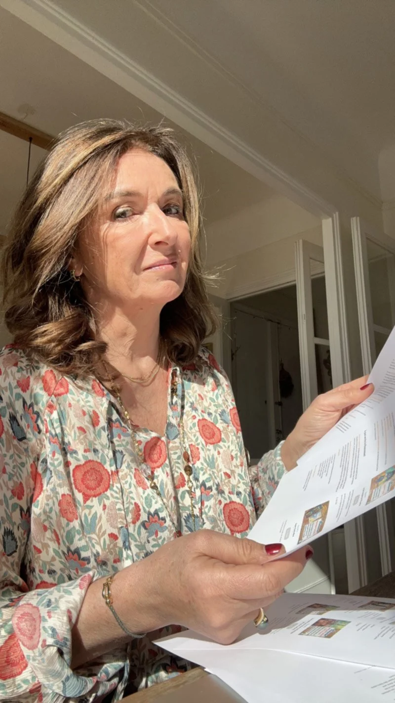 Une femme avec des cheveux bruns bouclés lit un document papier dans une pièce intérieure.