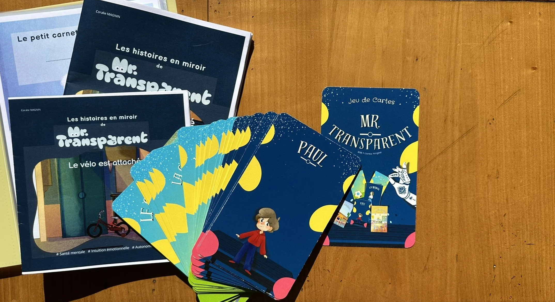 Jeu de cartes 'Mr Transparent' avec illustrations de personnages et éléments colorés, livres avec titres en français sur les histoires de Mr Transparent, le vélo attaché, tout sur une surface en bois.
