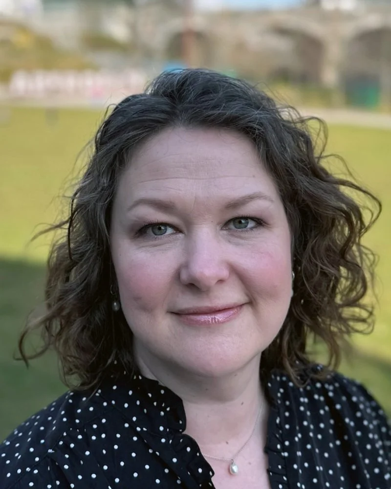 A woman with shoulder-length curly brown hair and blue eyes, wearing a black polka dot top and pearl earrings, smiling outdoors with a grassy field and buildings in the background.