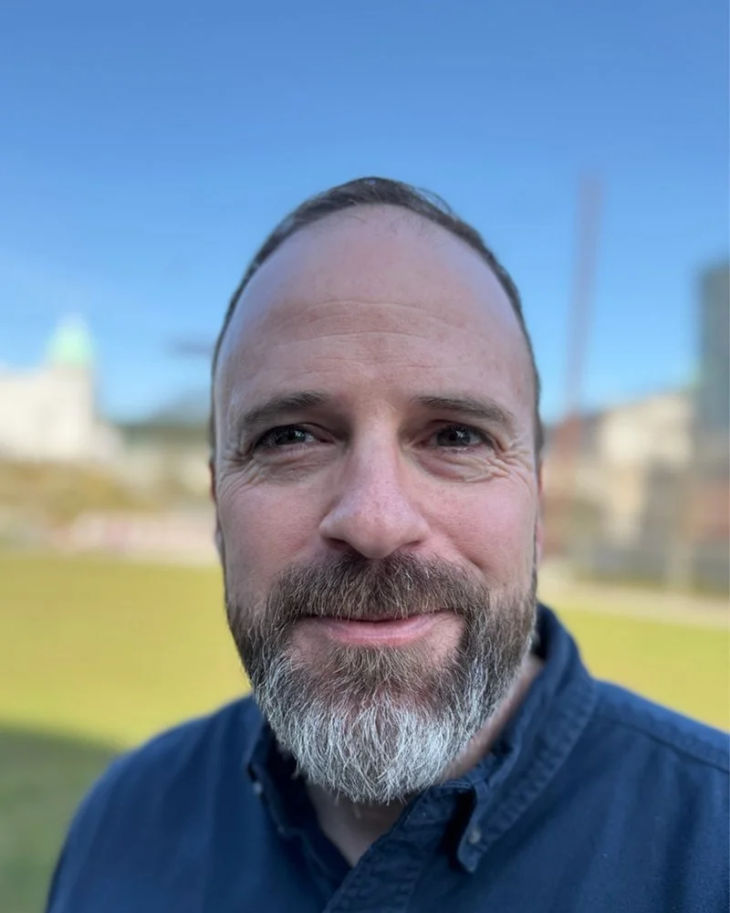 Close-up of a smiling man with a beard in an outdoor setting, with a clear blue sky in the background.