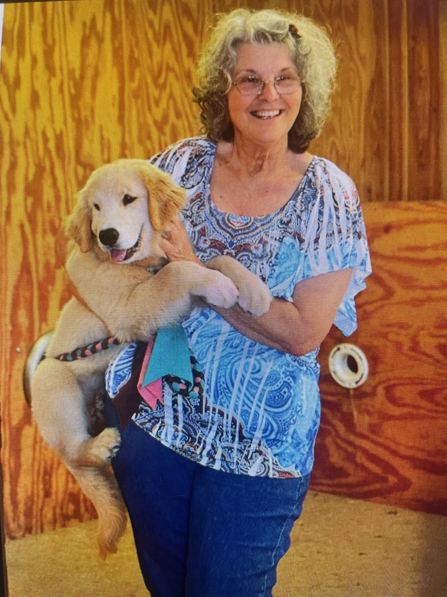 A woman with curly, gray hair and glasses smiling while holding a yellow Labrador retriever puppy with a birthday bandana sitting on her lap, in front of a wooden wall.