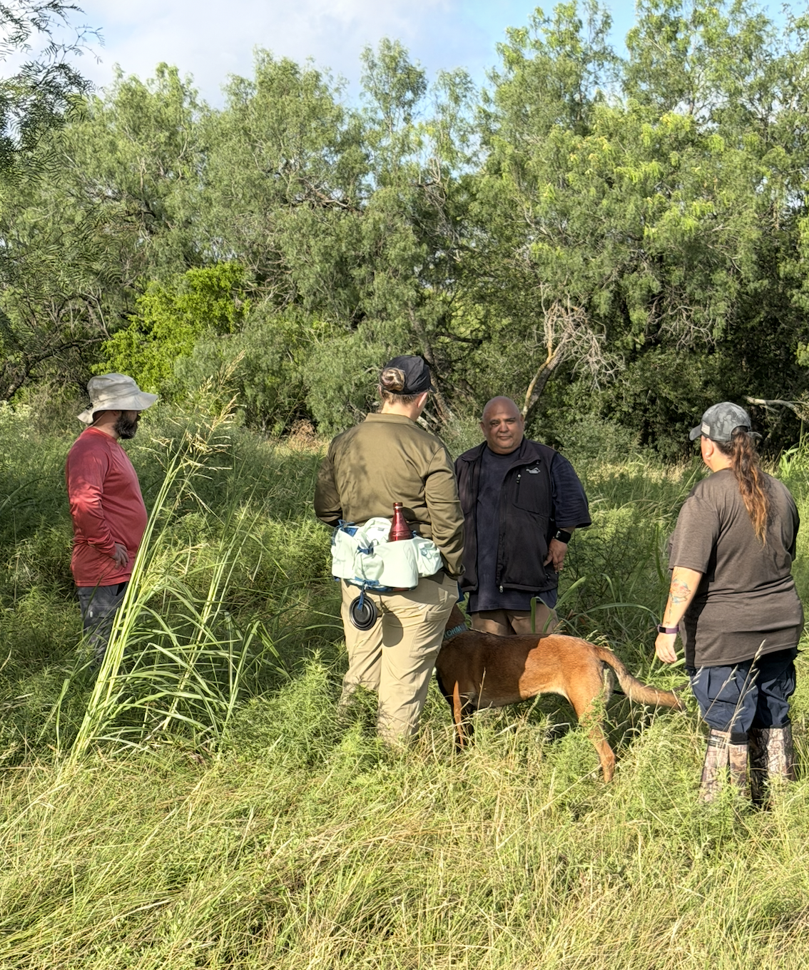 Four people and a dog standing in a grassy, wooded area having a conversation during scent and search training.