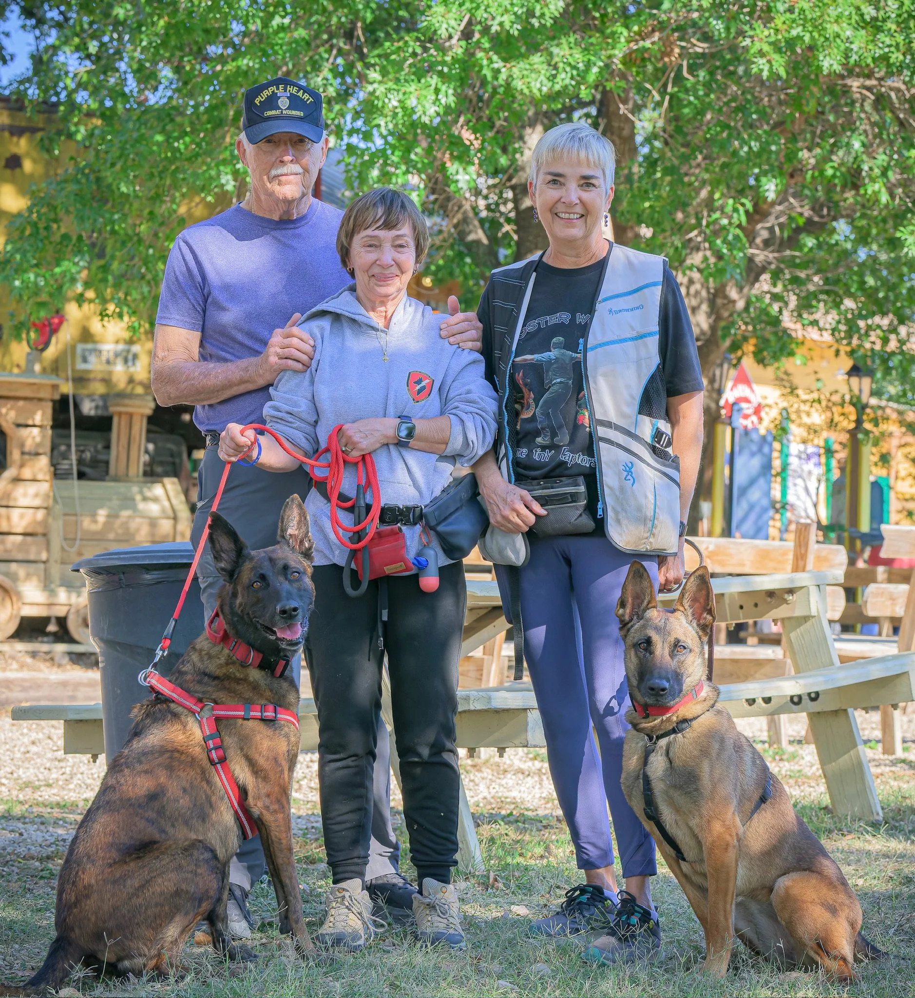 Three elderly people, two women and one man, stand outdoors in front of a large tree, with two German Shepherd dogs sitting nearby. The man wears a cap and casual clothing, the woman in the middle wears a light gray hoodie, and the woman on the right wears a vest and a black t-shirt. The scene appears to be in a park or a yard on a sunny day.