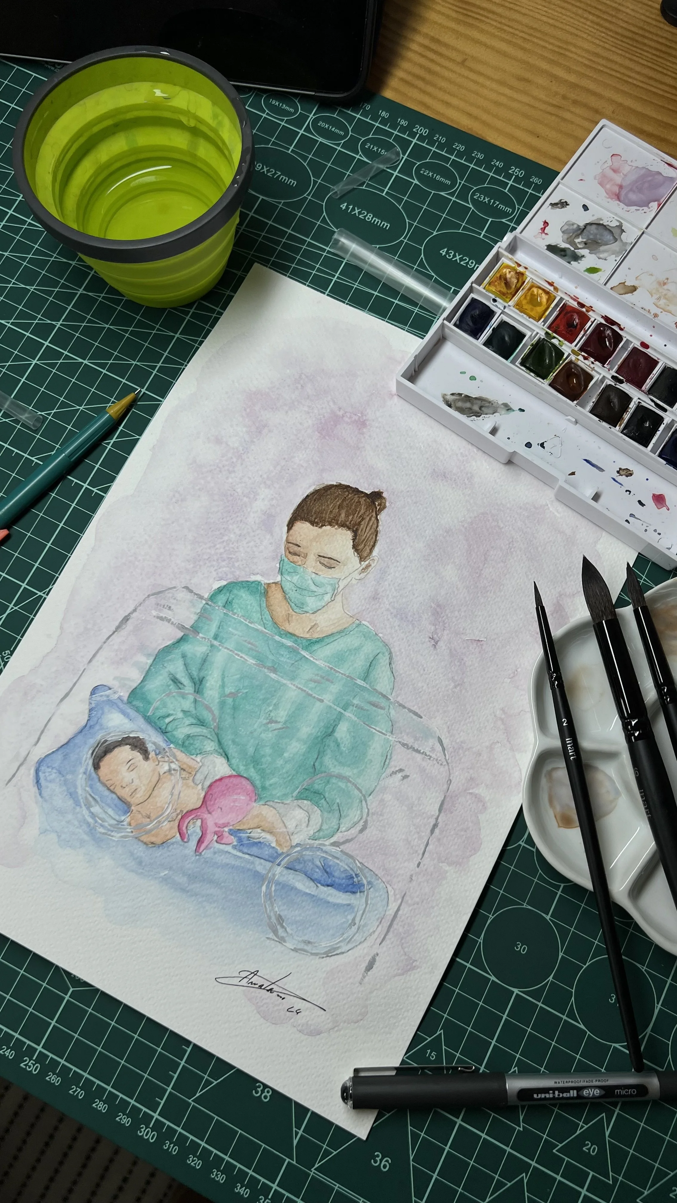 Mesa de arte com material de pintura aquarela, incluindo pincéis, paleta de tintas, copo com água, lápis, e uma folha de papel com a pintura de uma enfermeira com um bebê em uma incubadora.