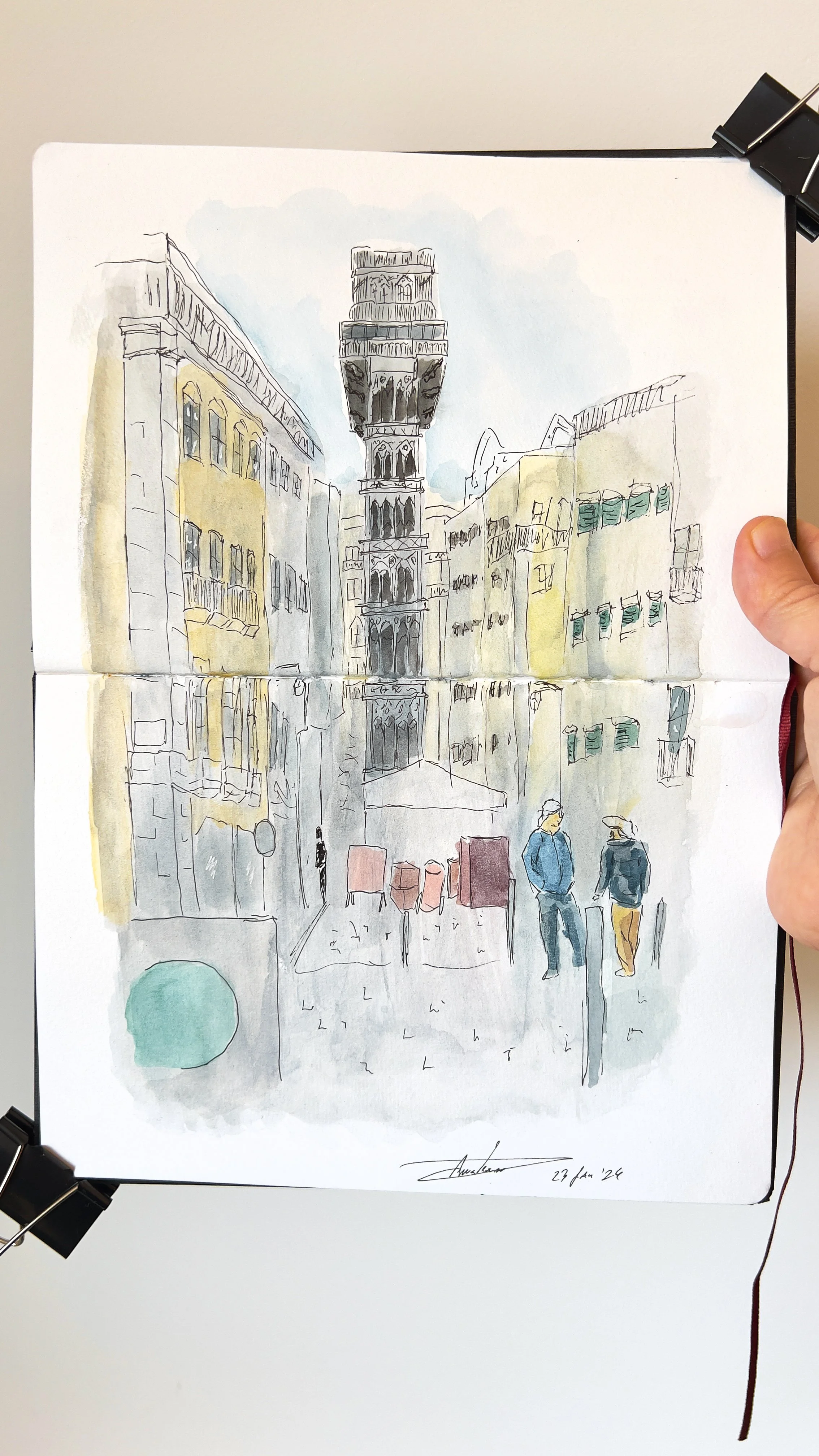 Desenho aquarela de uma rua estreita com prédios altos e uma torre no centro. Duas pessoas caminham na rua. O desenho está preso em um caderno com clipes pretos.