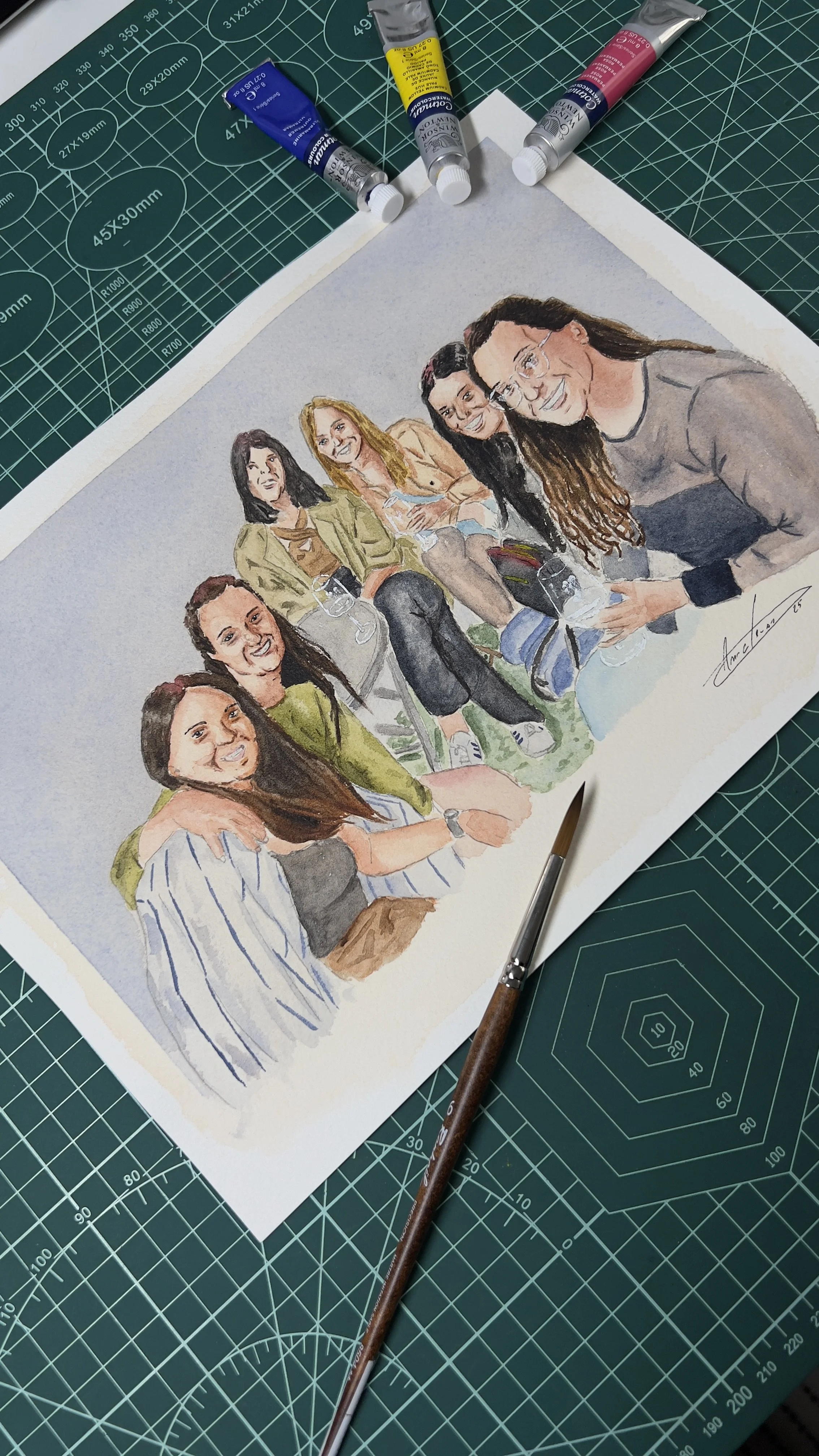 Uma pintura em aquarela de um grupo de pessoas sorrindo, cercada por tubos de tinta e um pincel sobre uma superfície com marcações de medidas.