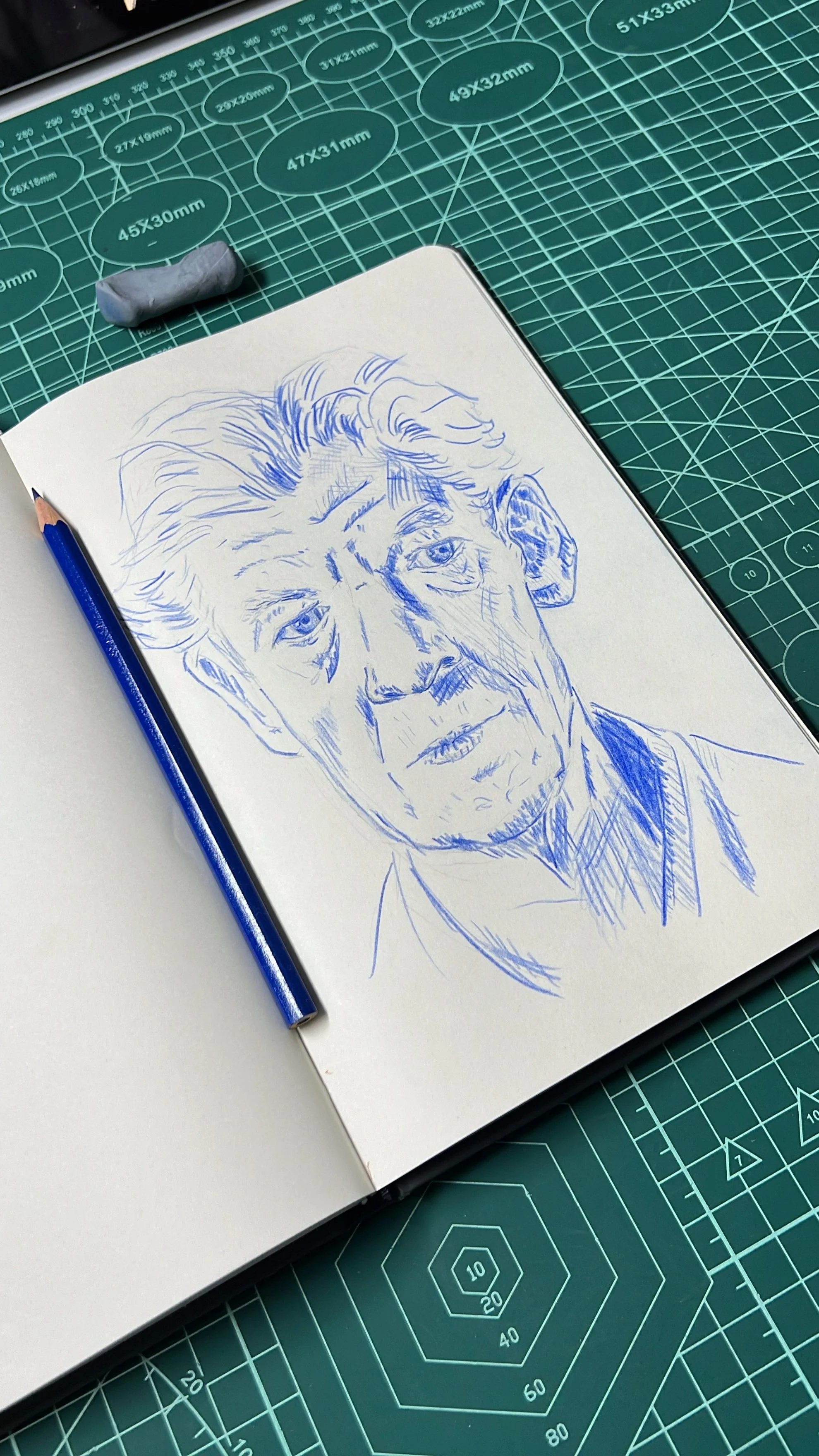 Retrato desenhado à mão em um caderno com lápis azul sobre uma mesa de corte verde.