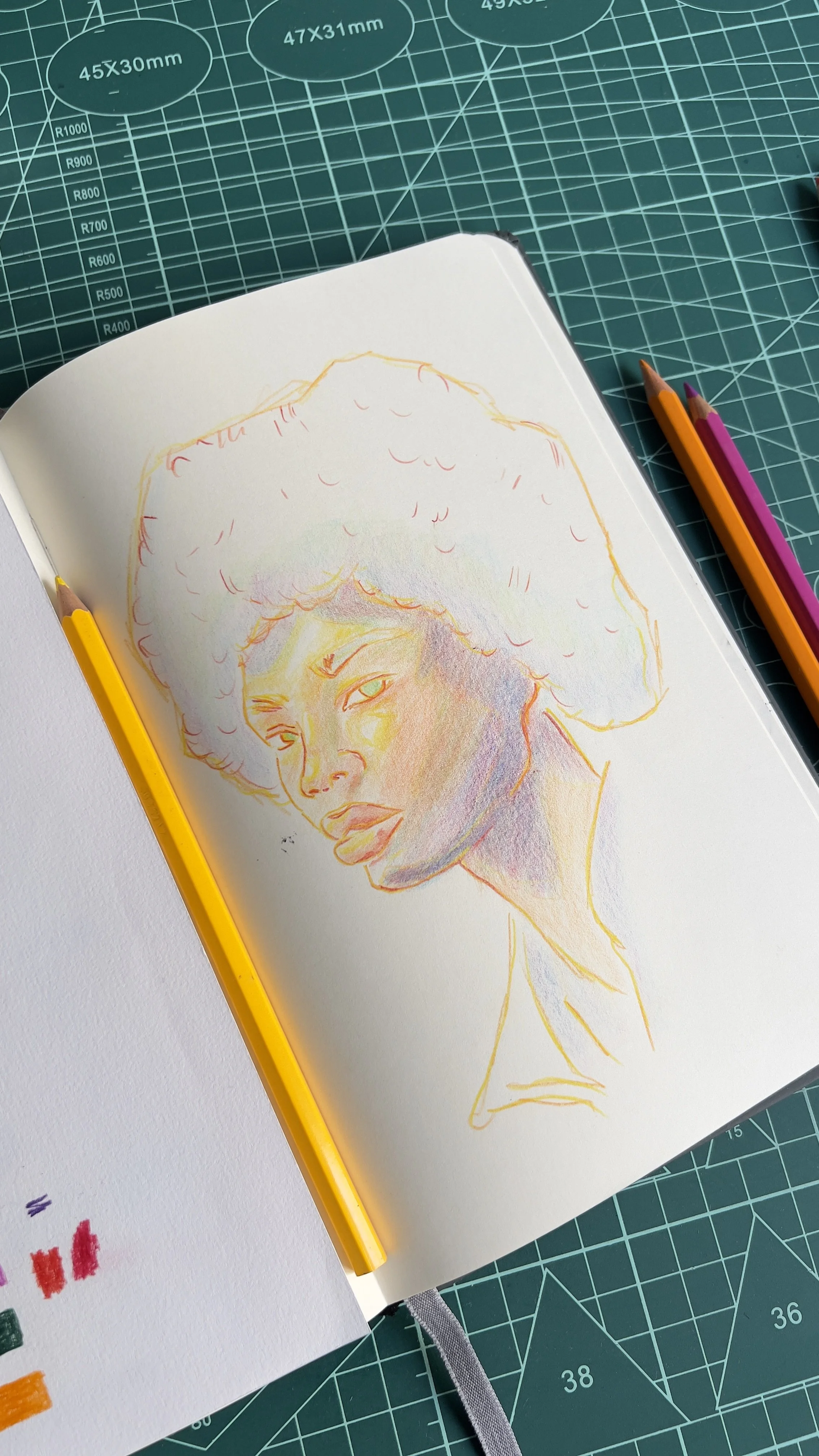 Desenho em caderno com linhas coloridas de uma pessoa de cabelo afro, com lápis de cor ao lado sobre uma mesa azul de corte.