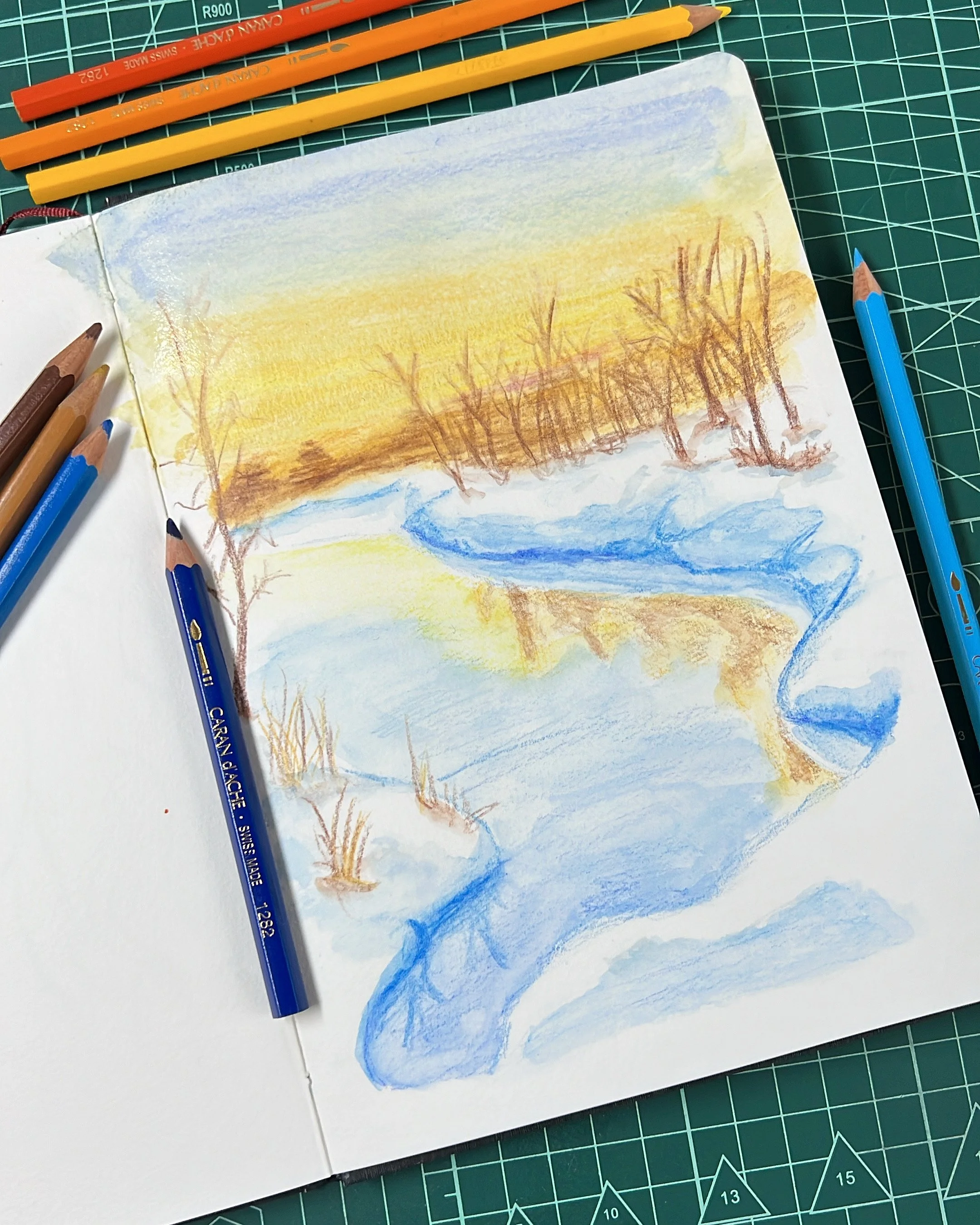 Desenho de paisagem de inverno em um caderno, com lápis de cor ao redor. A imagem mostra um rio sinuoso com margens cobertas de neve e árvores sem folhas ao fundo, sob um céu amarelo.