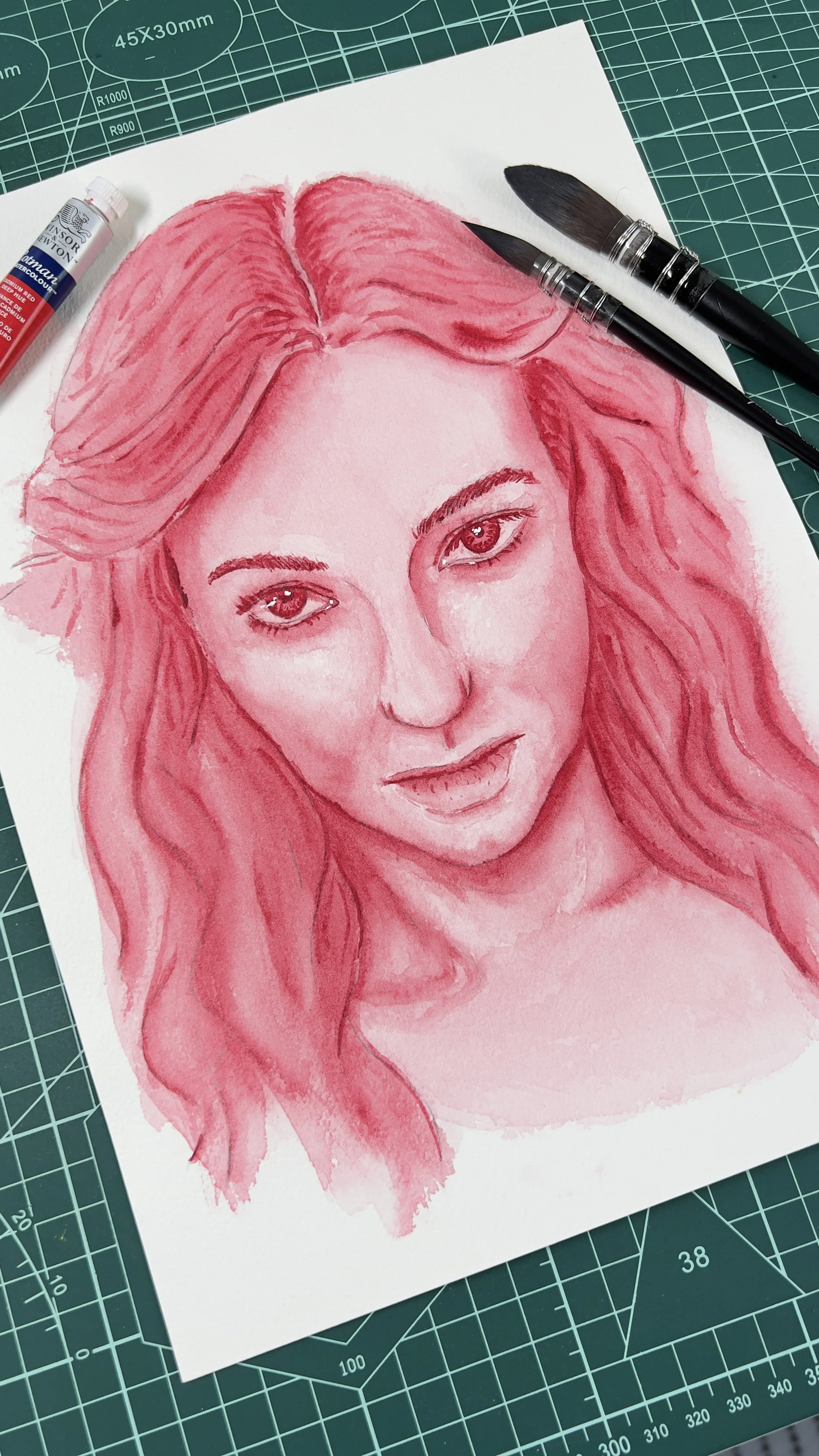 Retrato em aquarela de uma mulher em tons de rosa sobre uma mesa de corte. Ao lado, pincéis e um tubo de tinta. A mulher tem cabelos longos e expressão serena.