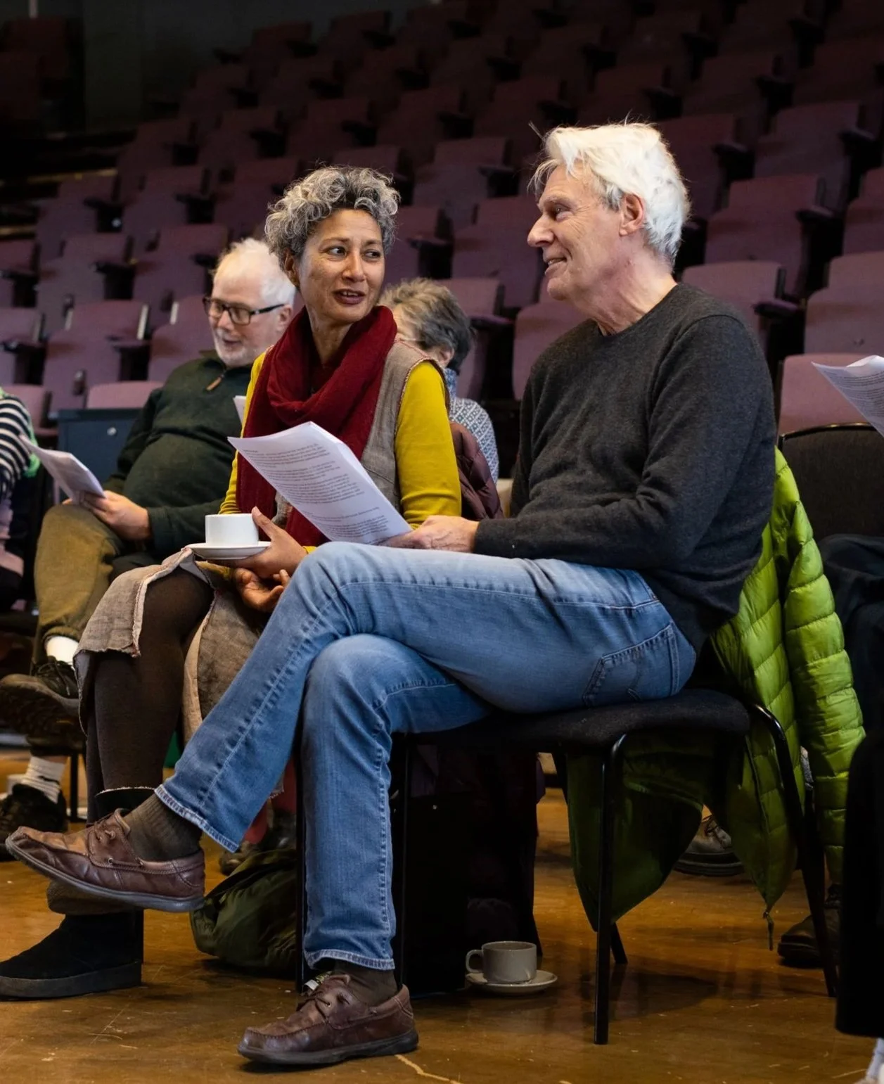 Group of people sitting in a row of chairs, engaged in conversation during what appears to be a meeting or workshop. The setting is an auditorium or conference room with purple seats and wooden floor.