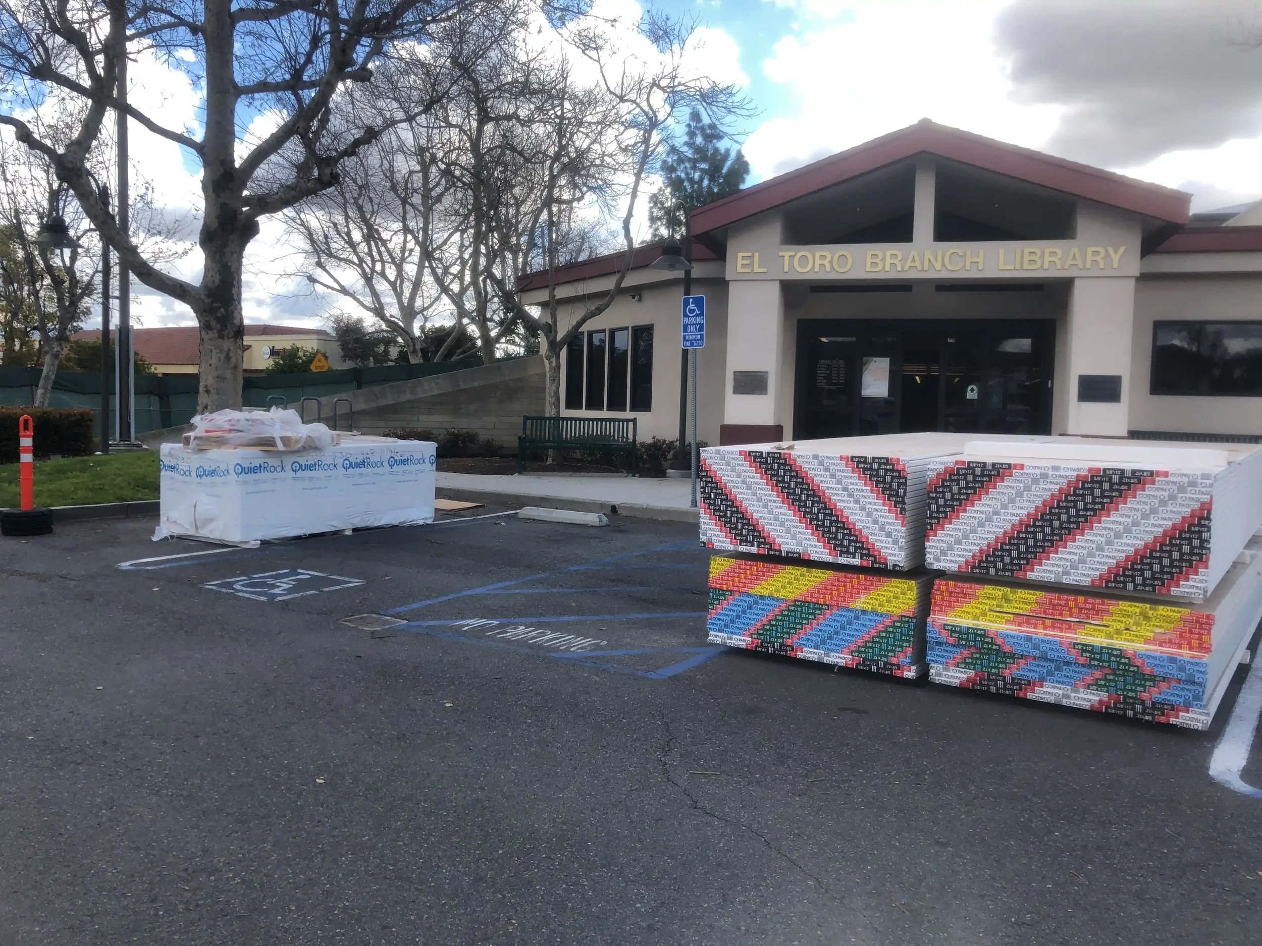 The exterior of El Toro Branch Library with a parking lot, leafless trees, and a cloudy sky. Outside, there are stacks of construction or promotional materials, including a white wrapped pallet and colorful patterned boxes.
