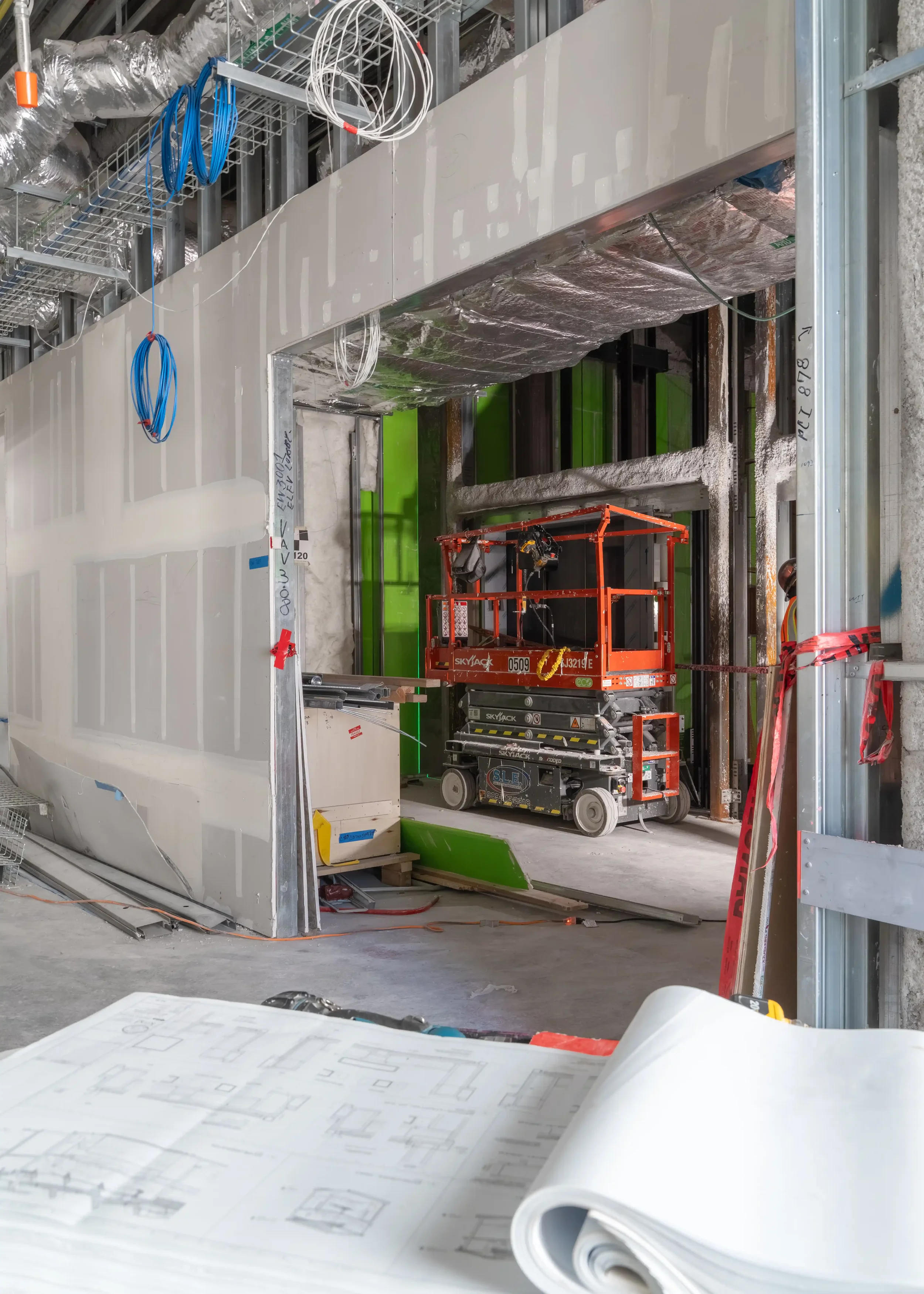 Construction site with an unfinished building, exposed metal framing, HVAC ductwork, wiring, and a suspended work platform. Blueprints and architectural plans are on a table in the foreground.