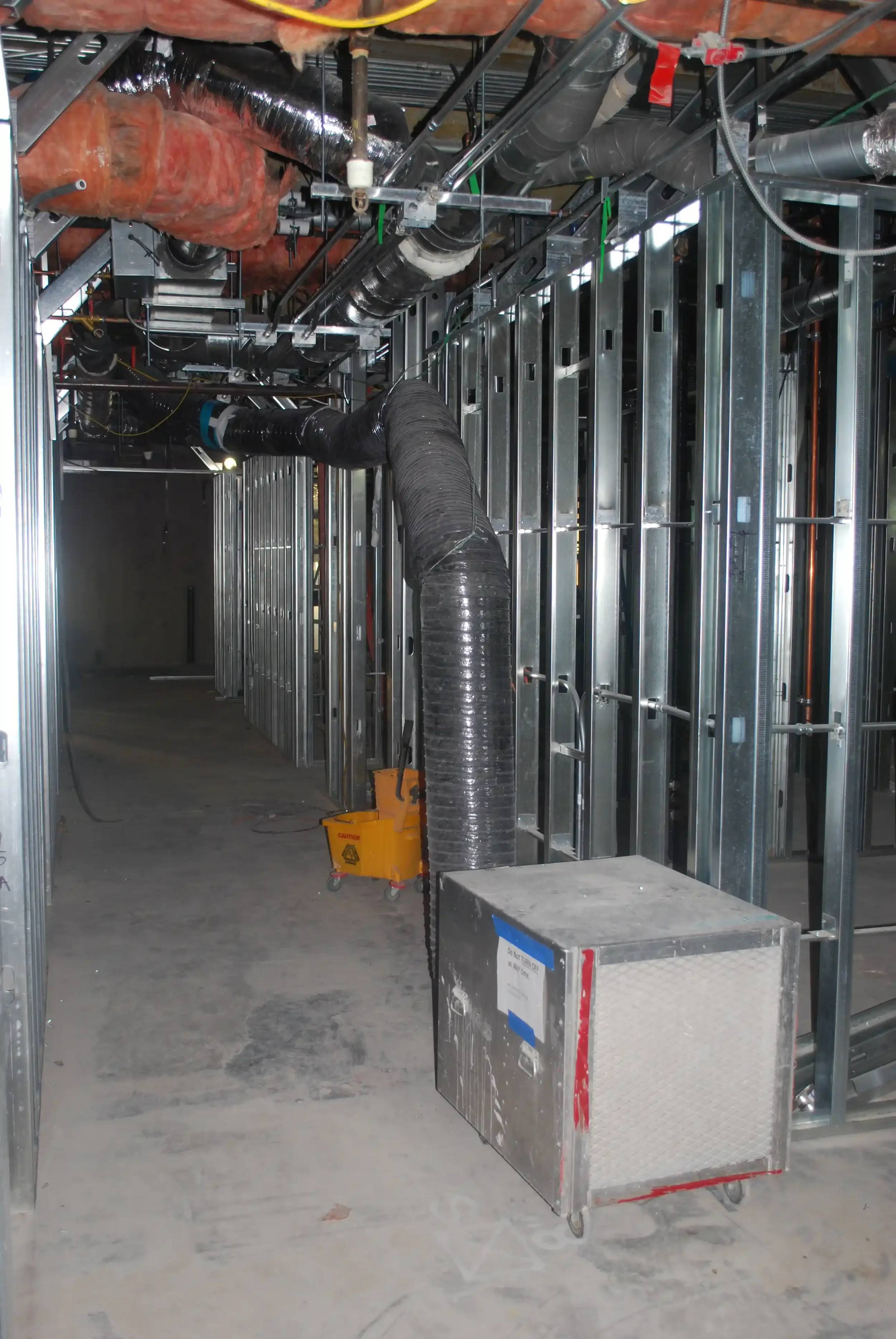 Construction site with metal framing, large black flexible ducts, orange insulation, and a yellow caution box on wheels.