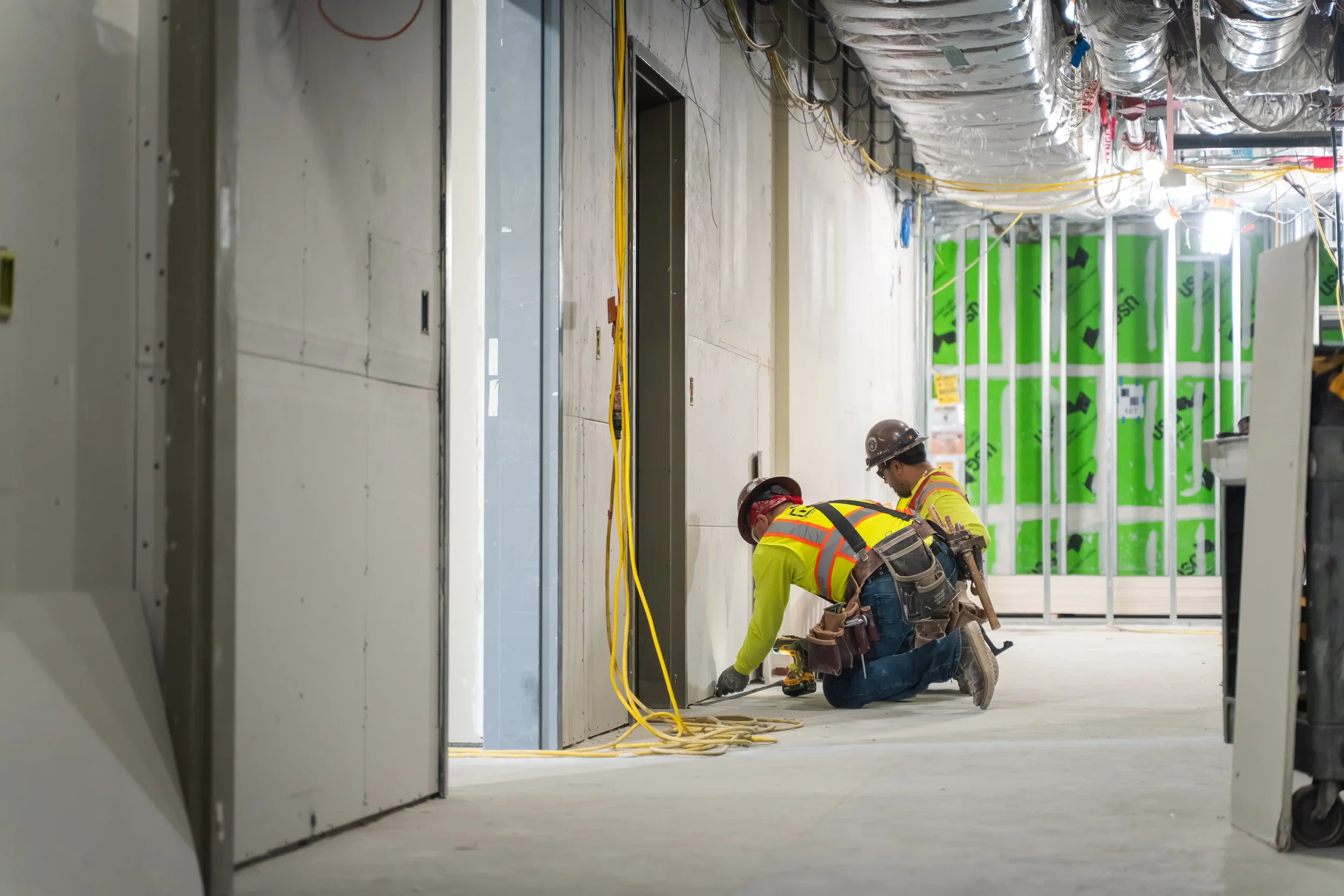 Two construction workers wearing yellow safety vests and hard hats, working on the floor of a building under construction. The room has exposed ducts and wiring, with green insulation on the walls.