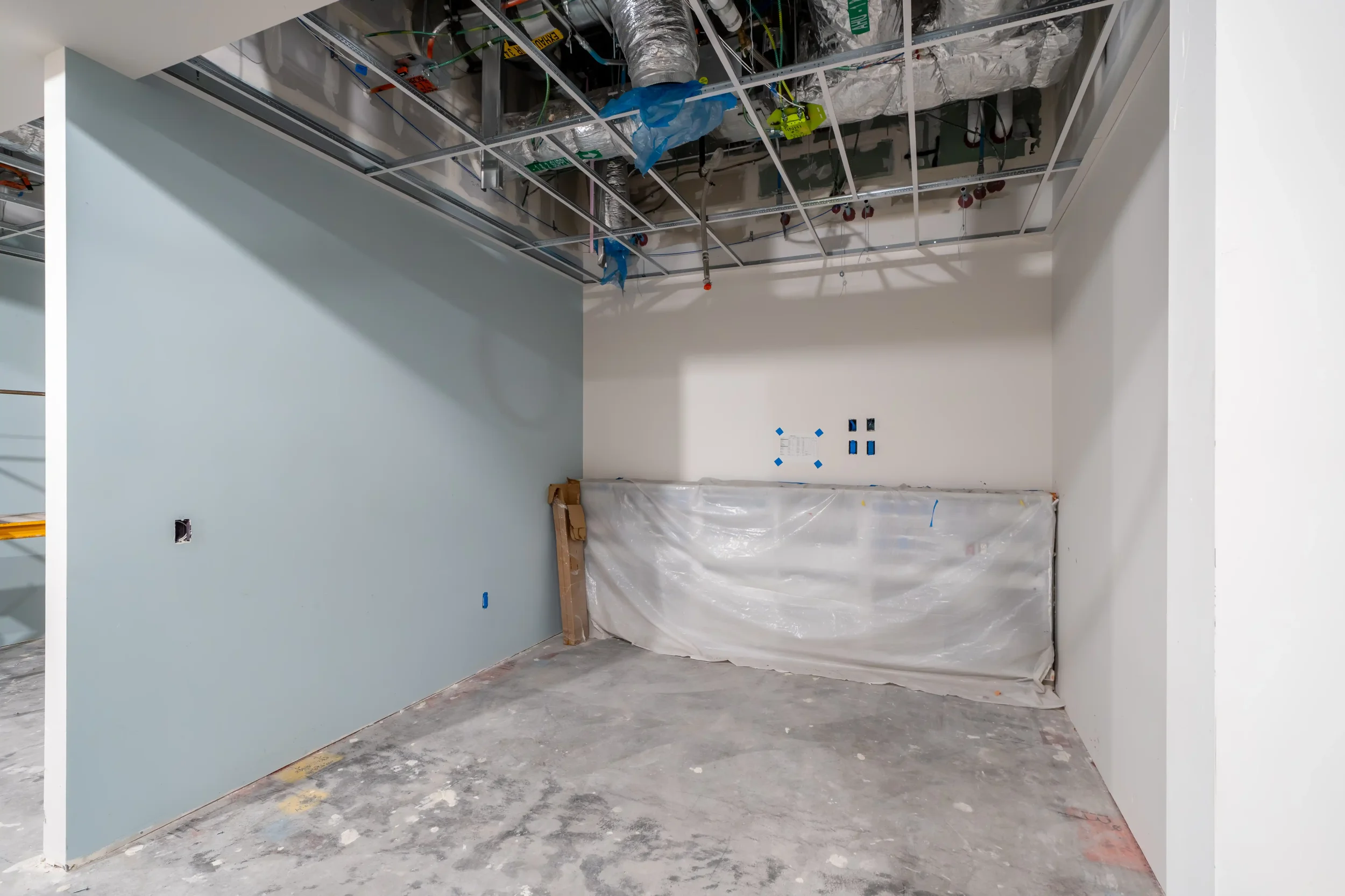 Construction area with exposed ceiling framework, ductwork, wiring, and partially painted walls. Plastic covering hanging, electrical outlets installed, and unfinished flooring.