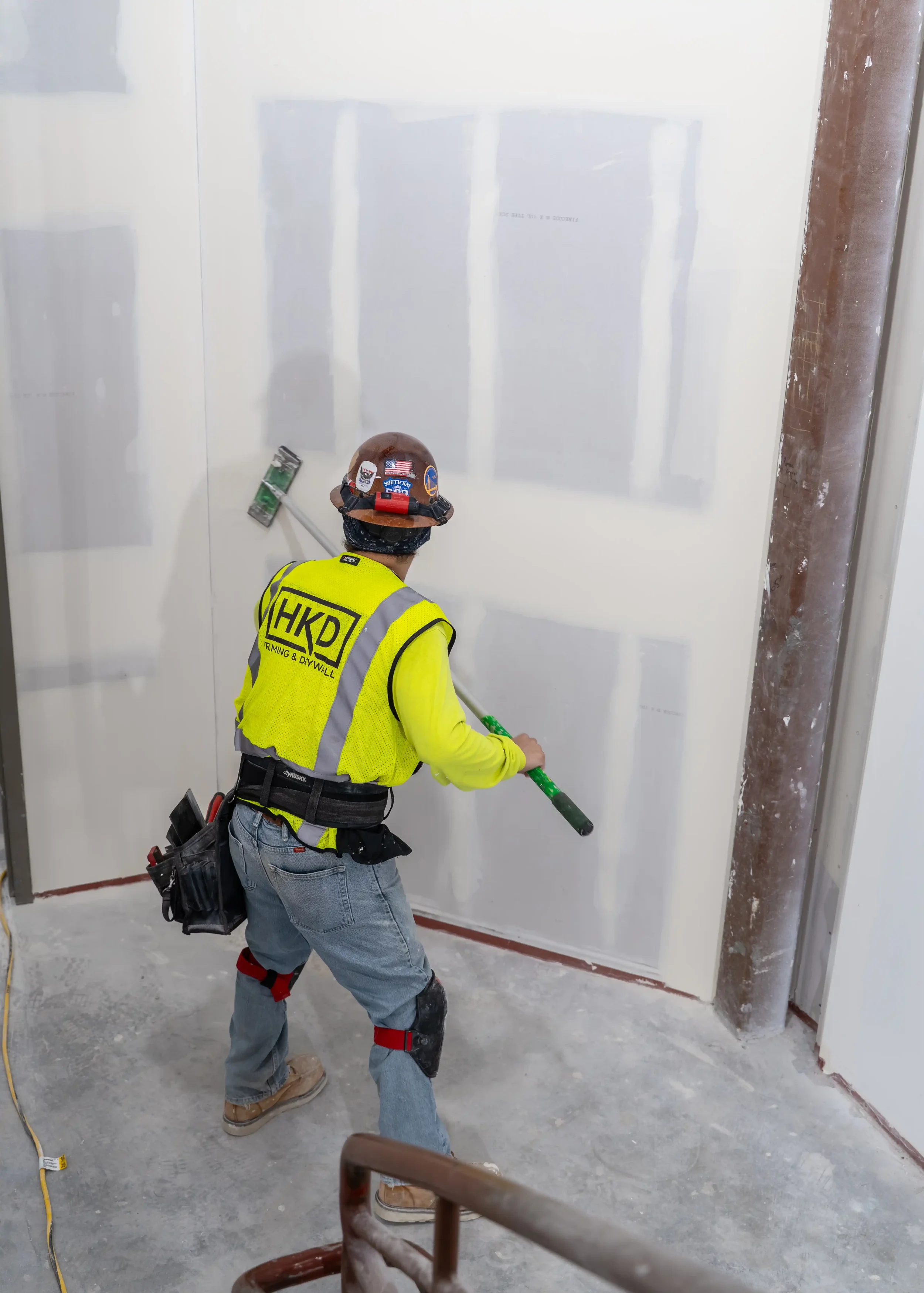 Construction worker in safety vest and helmet using a drywall sander on a wall inside a building under construction.