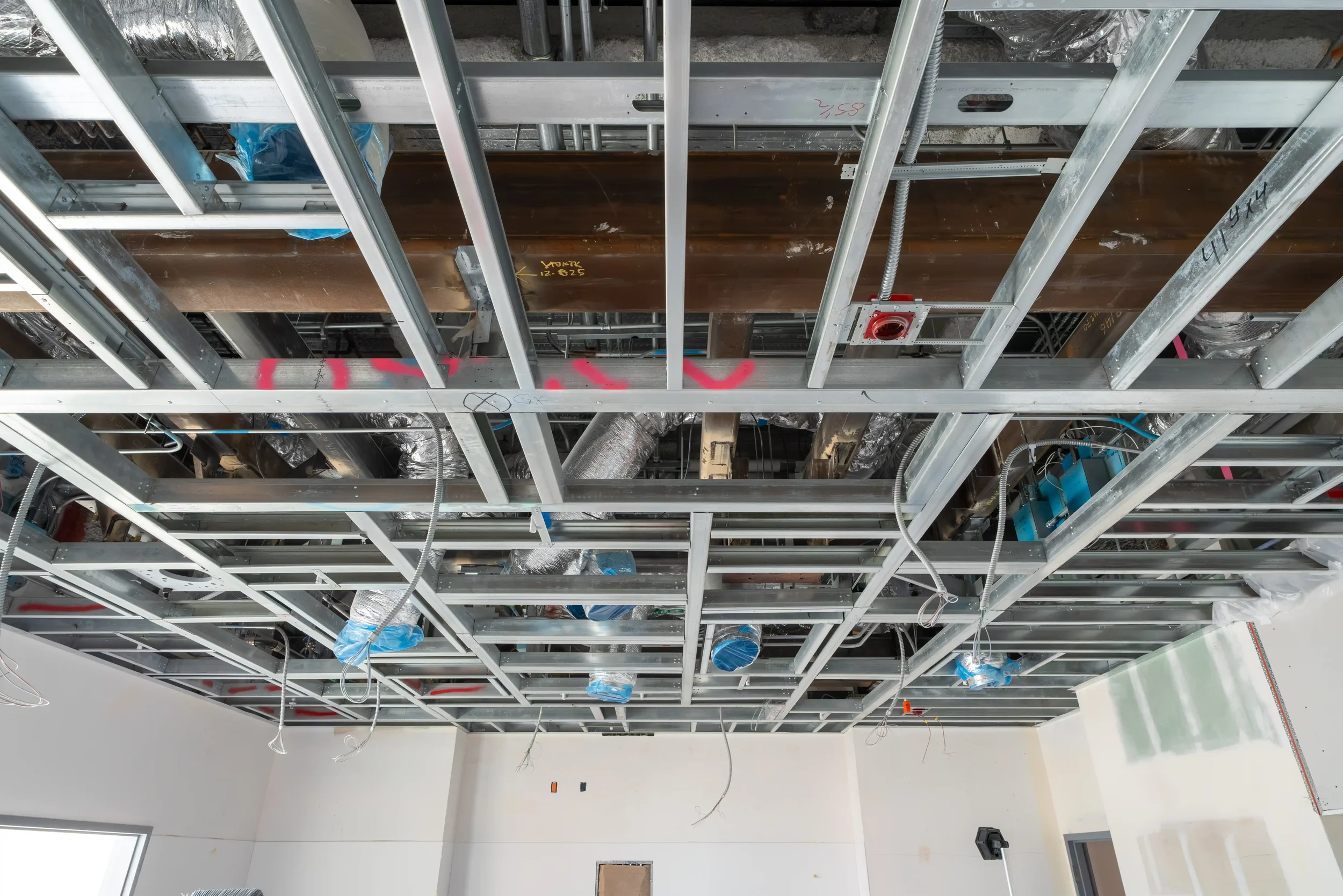 Construction ceiling framing with metal studs, mechanical ducts, wiring, and a red fire alarm call point.