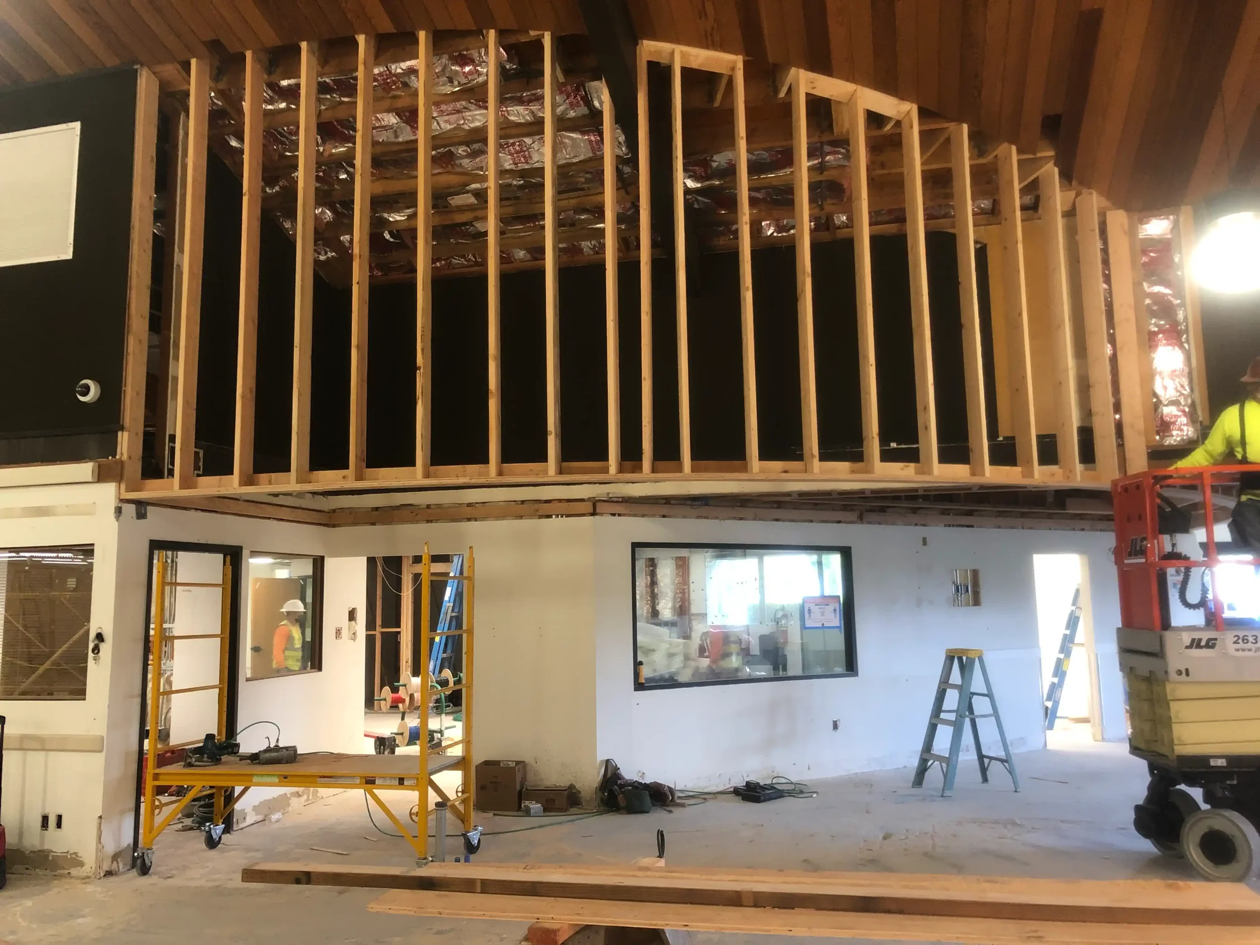Construction site interior with wooden framing and partially completed upper level. Tools, scaffolding, and construction workers are visible.