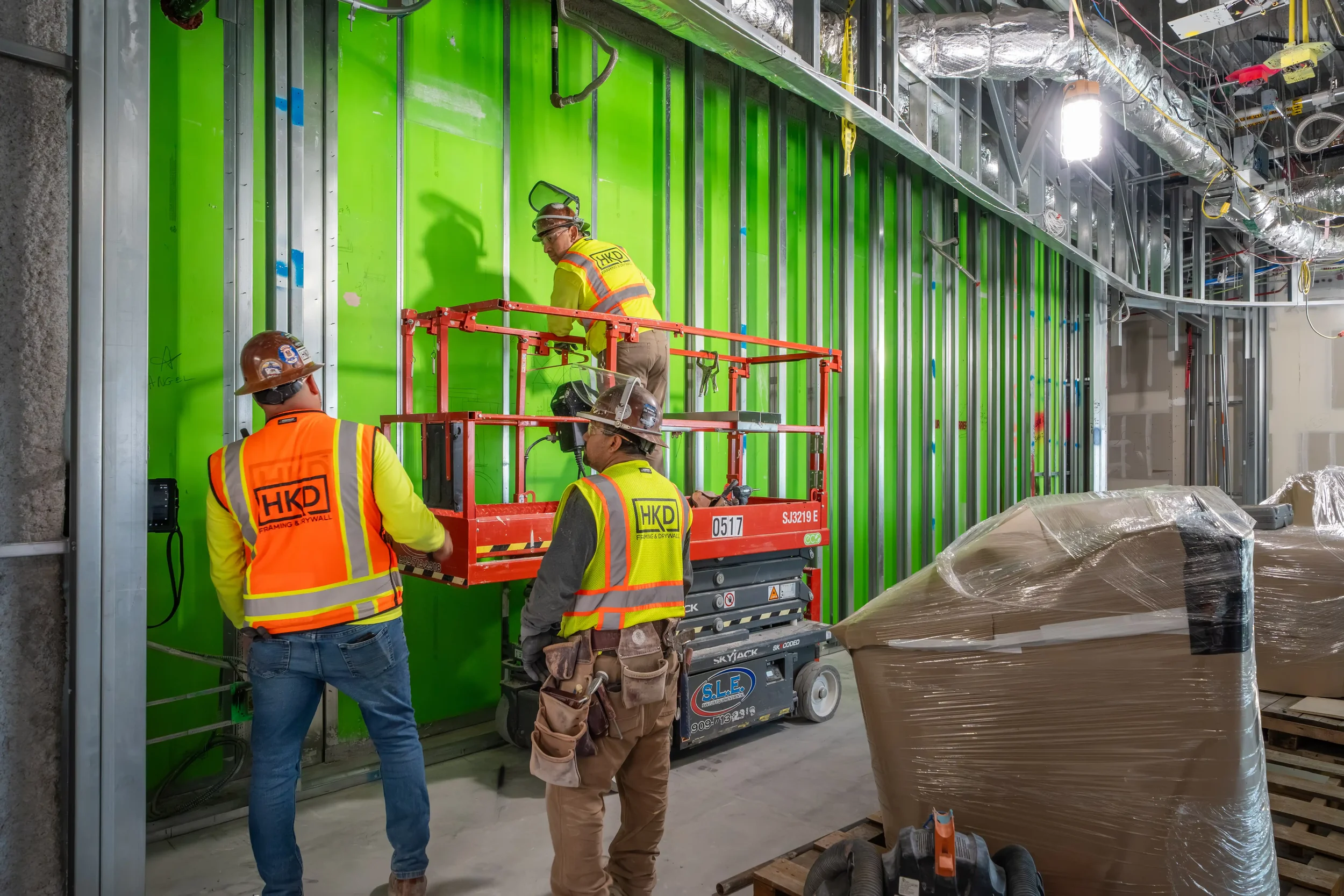 Construction workers installing framing and drywall in a building interior, with green protective wall panels and construction tools and equipment around.