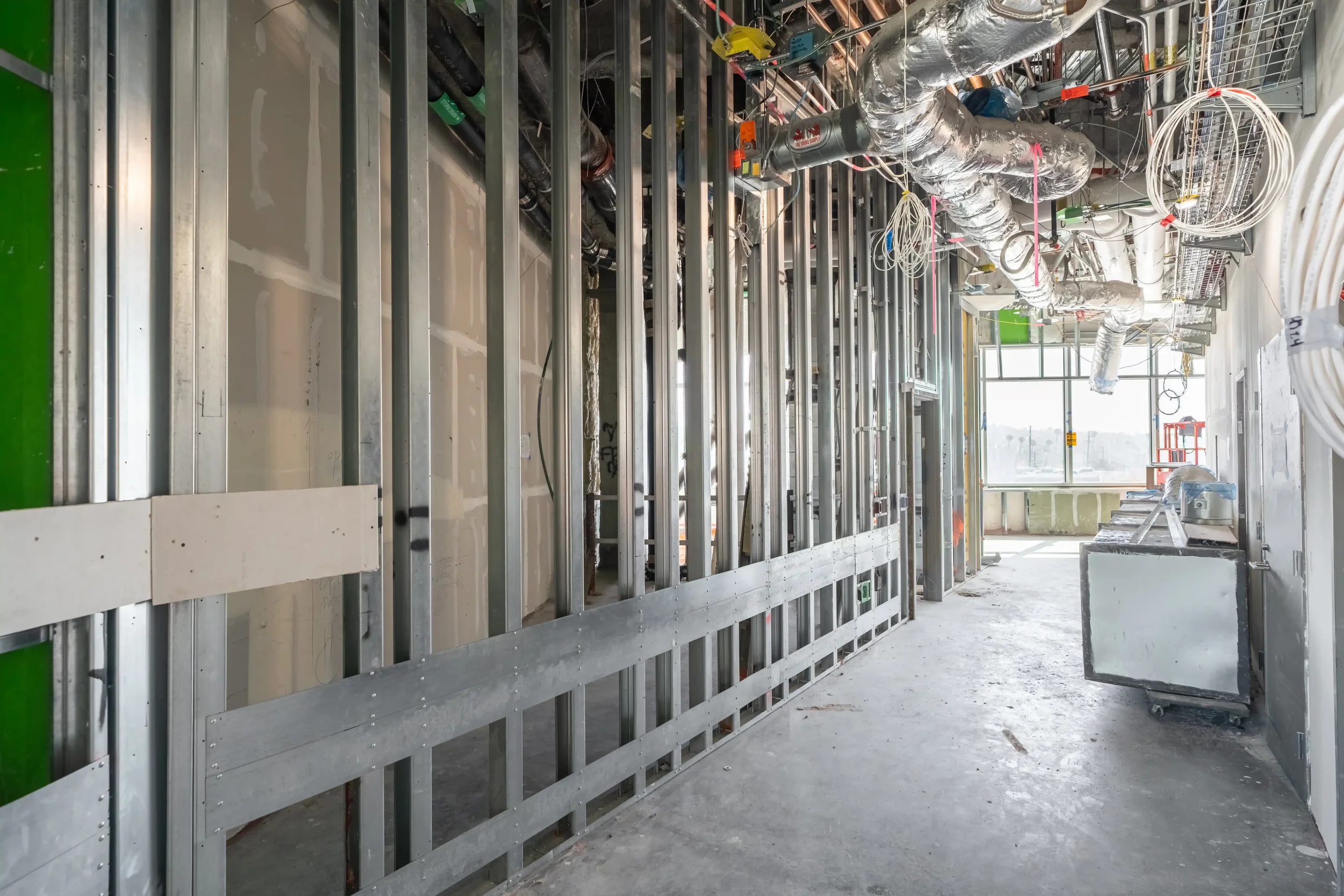 Interior view of a building under construction showing metal framing, electrical wiring, and HVAC ductwork in a large open space with windows at the far end.