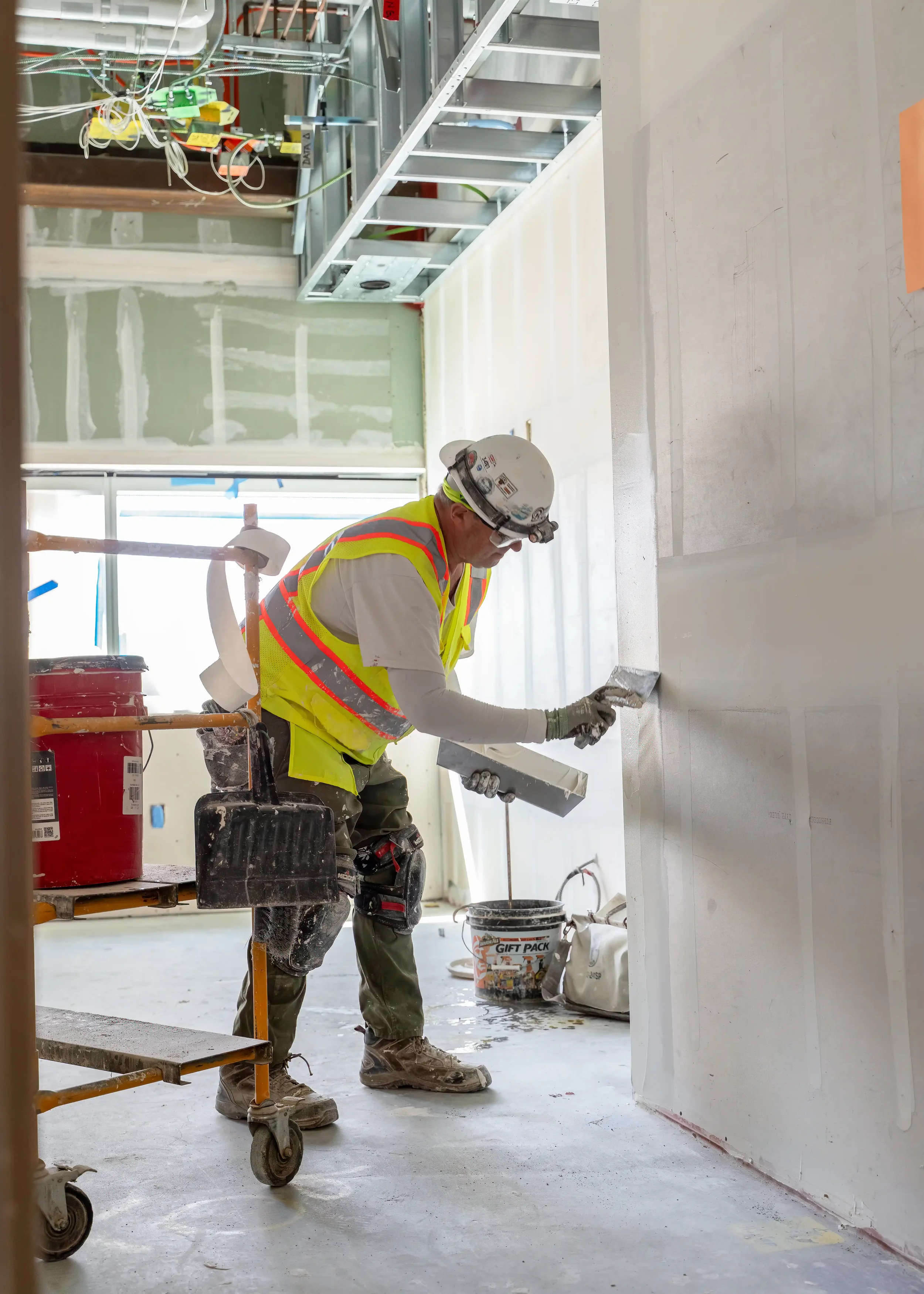 Construction worker in a hard hat and safety vest smoothing wet concrete inside a building under construction, with scaffolding, tools, and construction materials visible around him.